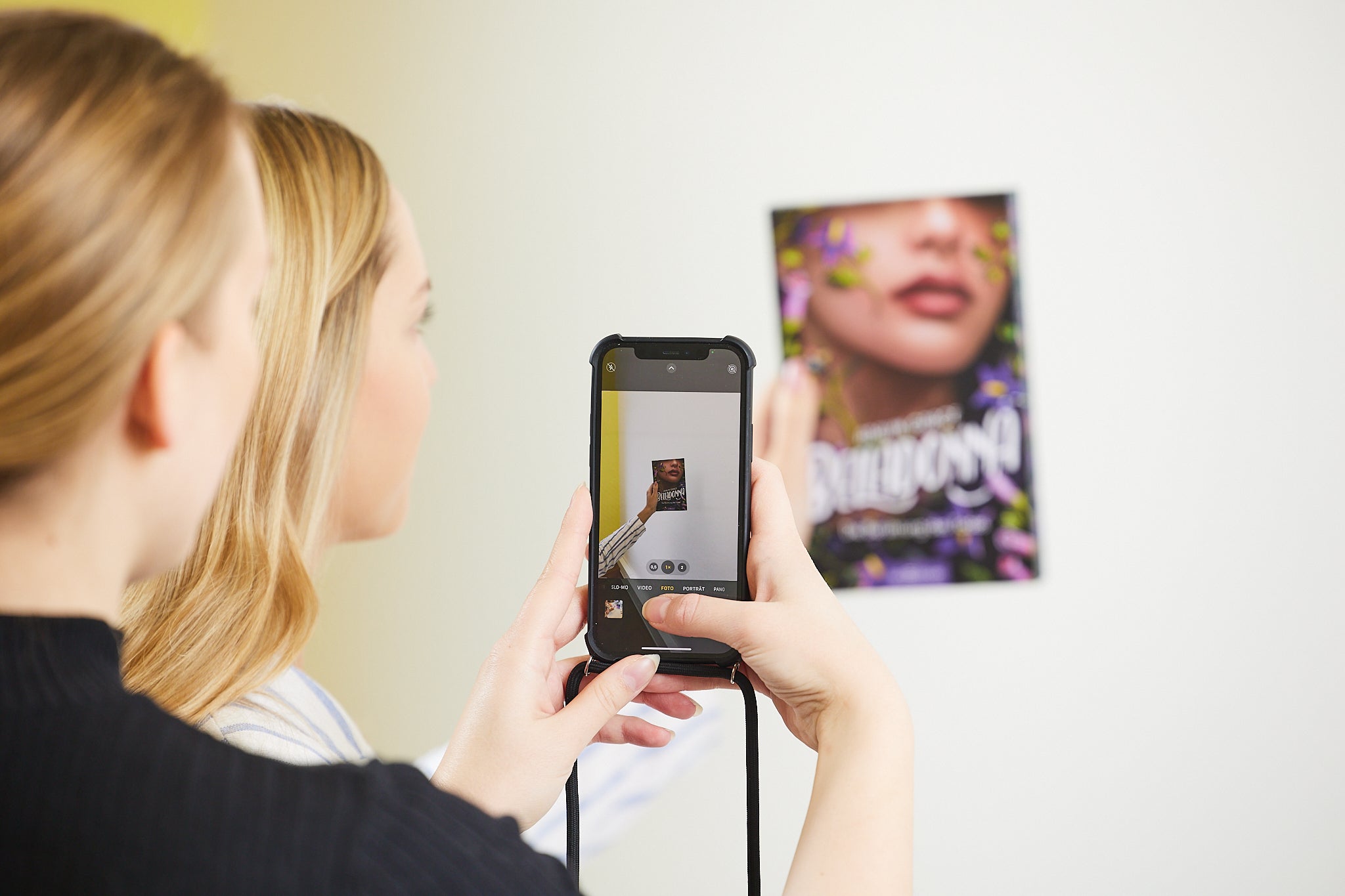Zwei Personen stehen vor einem bunten Plakat an der Wand. Eine Person hält ein Smartphone in der Hand und fotografiert das Buch, das auf dem Bildschirm des Smartphones klar zu erkennen ist. Der Hintergrund ist schlicht und hell.