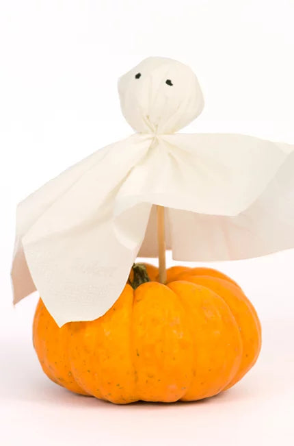 Ein kleines, orangefarbenes Kürbis-Dekor mit einer niedlichen Halloween-Figur darauf: Die Figur besteht aus einem weißen Taschentuch, das wie ein Geist gestaltet ist, mit aufgemalten schwarzen Augen und auf einem Holzstab befestigt.