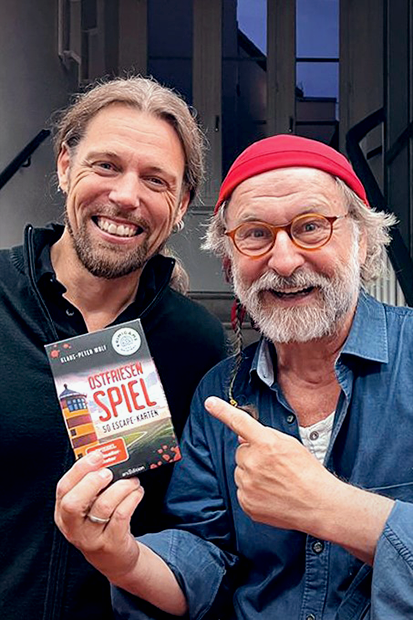 Gezeigt wird eine Innen- oder Zusatzansicht von „Ostfriesenspiel Sonderausgabe mit Autogrammkarte“. Die Abbildung zeigt zwei lächelnde Männer, die gemeinsam das Spiel „Ostfriesen-Spiel“ präsentieren, das sich in der linken Hand eines der Männer befindet. Das Cover des Spiels ist farbenfroh gestaltet, mit einer dominierenden Farbpalette aus Blau, Rot und Gelb. Die Schriftart ist modern und gut lesbar, der Titel hebt sich klar vom Hintergrund ab. Im Hintergrund ist eine helle,...