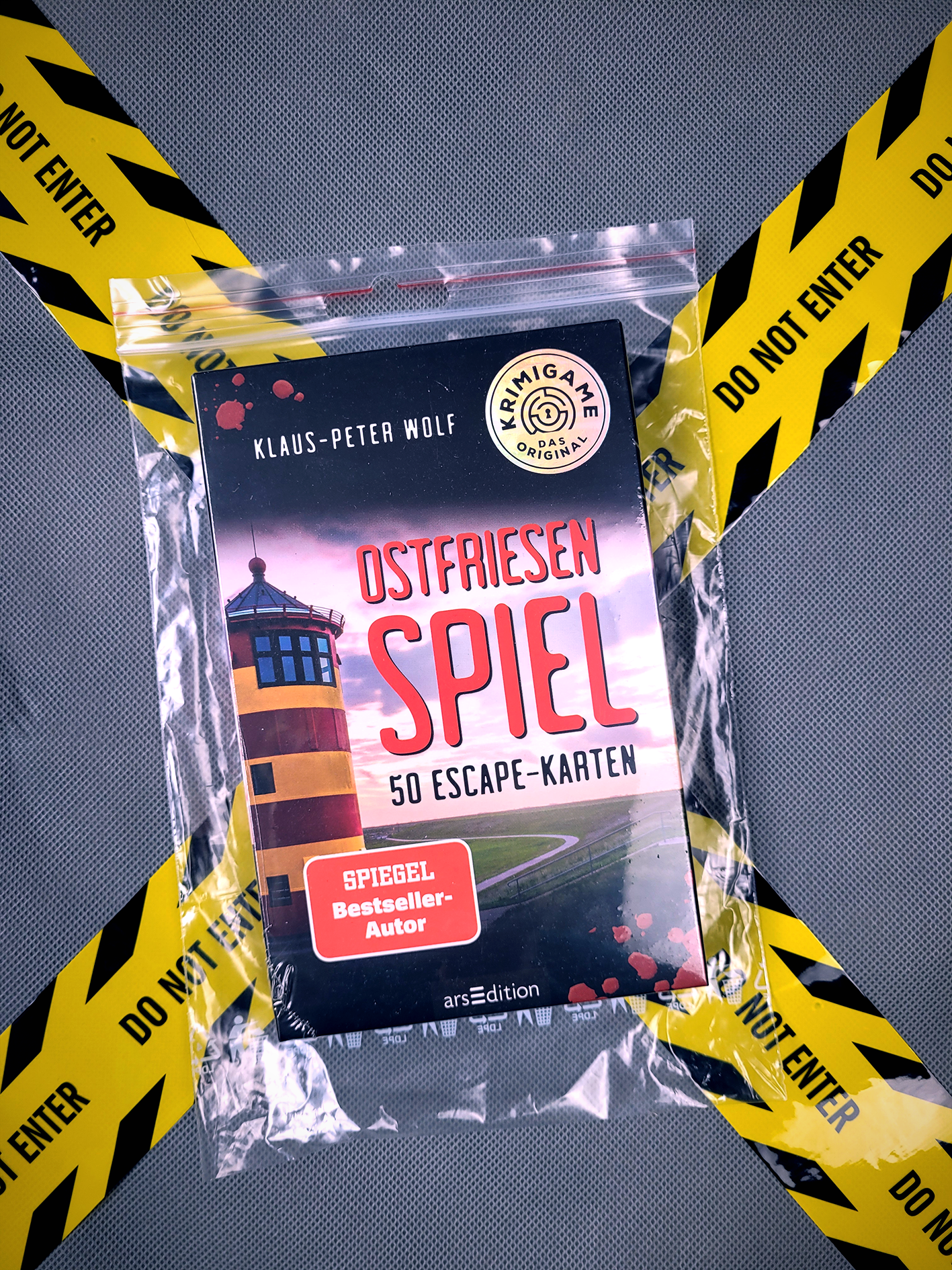 Das Buchcover zeigt eine stilisierte Darstellung eines Leuchtturms in leuchtenden Gelb- und Schwarztönen, isodromische Linien und eine kurvige Straßenführung, die den Hintergrund bildet. Der Titel „OSTFRIESEN SPIEL“ erscheint in großen, weißen Buchstaben und ist zentral platziert. Oben links befindet sich der Name des Autors Klaus-Peter Wolf. Ein rotes Banner mit dem Text „SPIEGEL Bestseller-Autor“ hebt den Erfolg des Titels hervor. Ganz unten mittig steht der Logoschriftzug von...