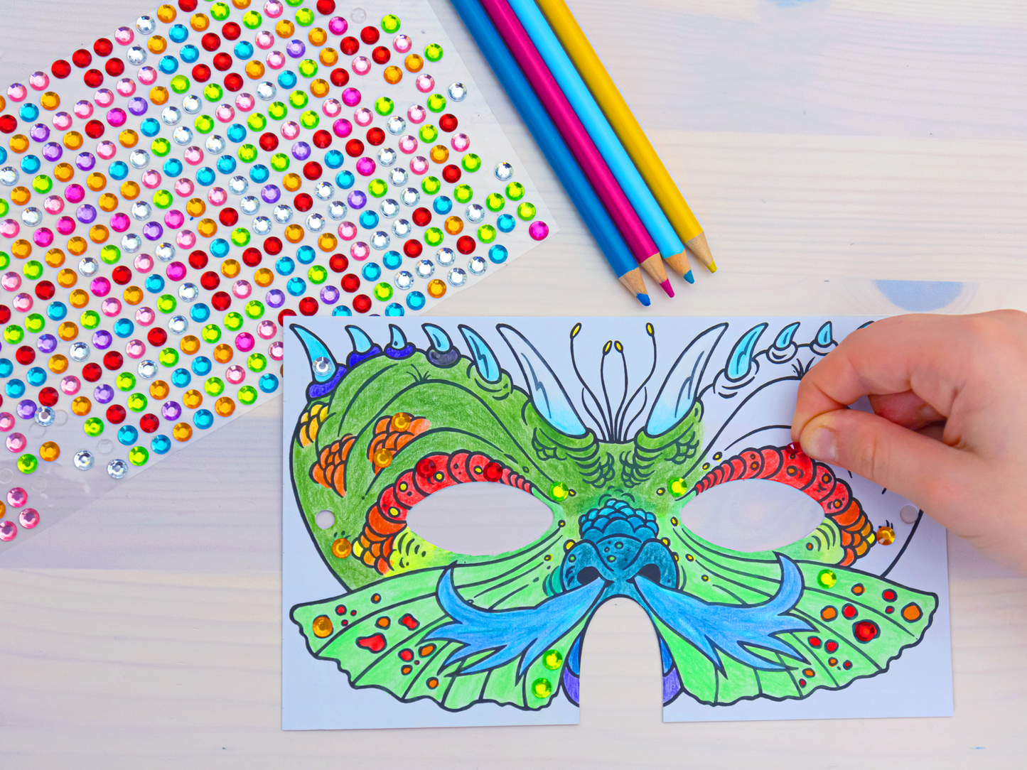 Gezeigt wird eine Innen- oder Zusatzansicht von „Diamantmasken – Zauberwesen“. Die Abbildung zeigt eine detaillierte, farbenfrohe Maskenzeichnung in Grüntönen sowie Akzenten in Blau und Rot. Neben der Maske befinden sich zahlreiche bunte, glitzernde Steinchen und ein Set Buntstifte, die ein kreatives Bastelerlebnis fördern. Die Typografie ist verspielt und ansprechend, während die insgesamt lebendige Gestaltung die fantasievolle Atmosphäre betont.