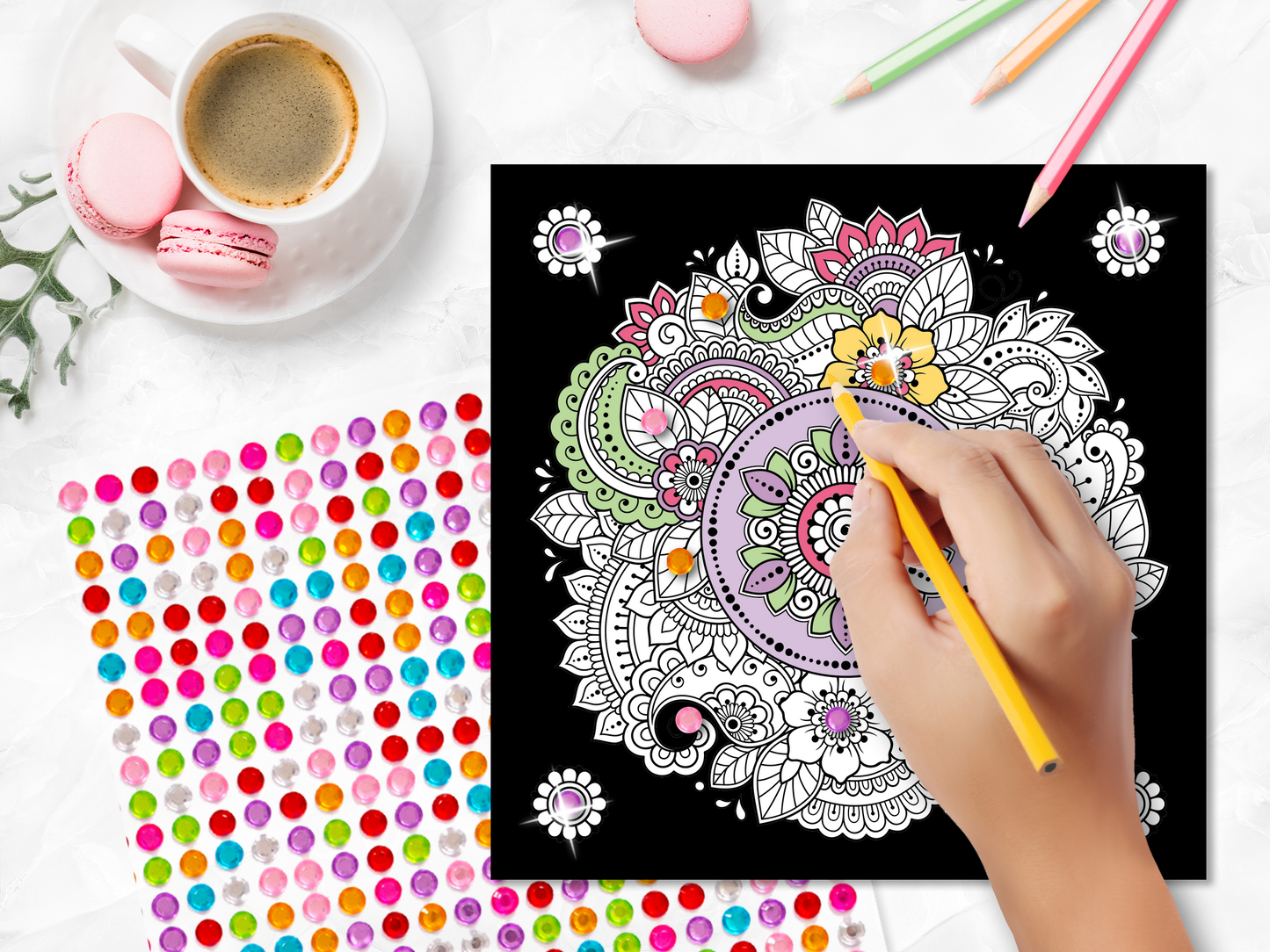 Gezeigt wird eine Innen- oder Zusatzansicht von „Diamond Mandalas“. Im Vordergrund ist eine bunte Mandala-Zeichnung auf schwarzem Hintergrund zu sehen, die mit verschiedenen Farben, darunter gelb, grün und rosa, verziert ist. Eine Hand hält einen gelben Stift und färbt die Zeichnung aus. Zudem sind bunte Dekosticker und pastellfarbene Macarons auf einem weißen Tisch sichtbar, sowie eine Tasse Kaffee im Hintergrund. DieTypografie wird durch stilisierte Schriftzüge ergänzt.