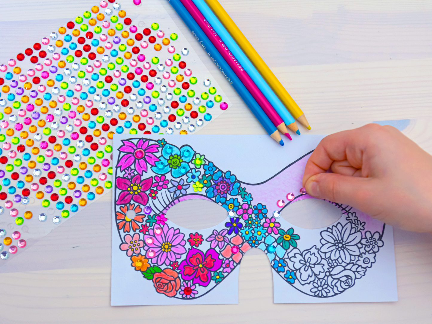 Gezeigt wird eine Innen- oder Zusatzansicht von „Diamantmasken – Feen“. Die Abbildung zeigt eine bunte, handgezeichnete Maske im Mittelpunkt, verziert mit floralen Motiven in lebendigen Farben wie Pink, Blau und Gelb. Um die Maske herum liegen bunte Buntstifte und ein Blatt mit glänzenden, klebenden Strasssteinen in verschiedenen Farben. Eine Hand platziert vorsichtig einen Strassstein auf der Maske, was den kreativen Gestaltungsprozess verdeutlicht.