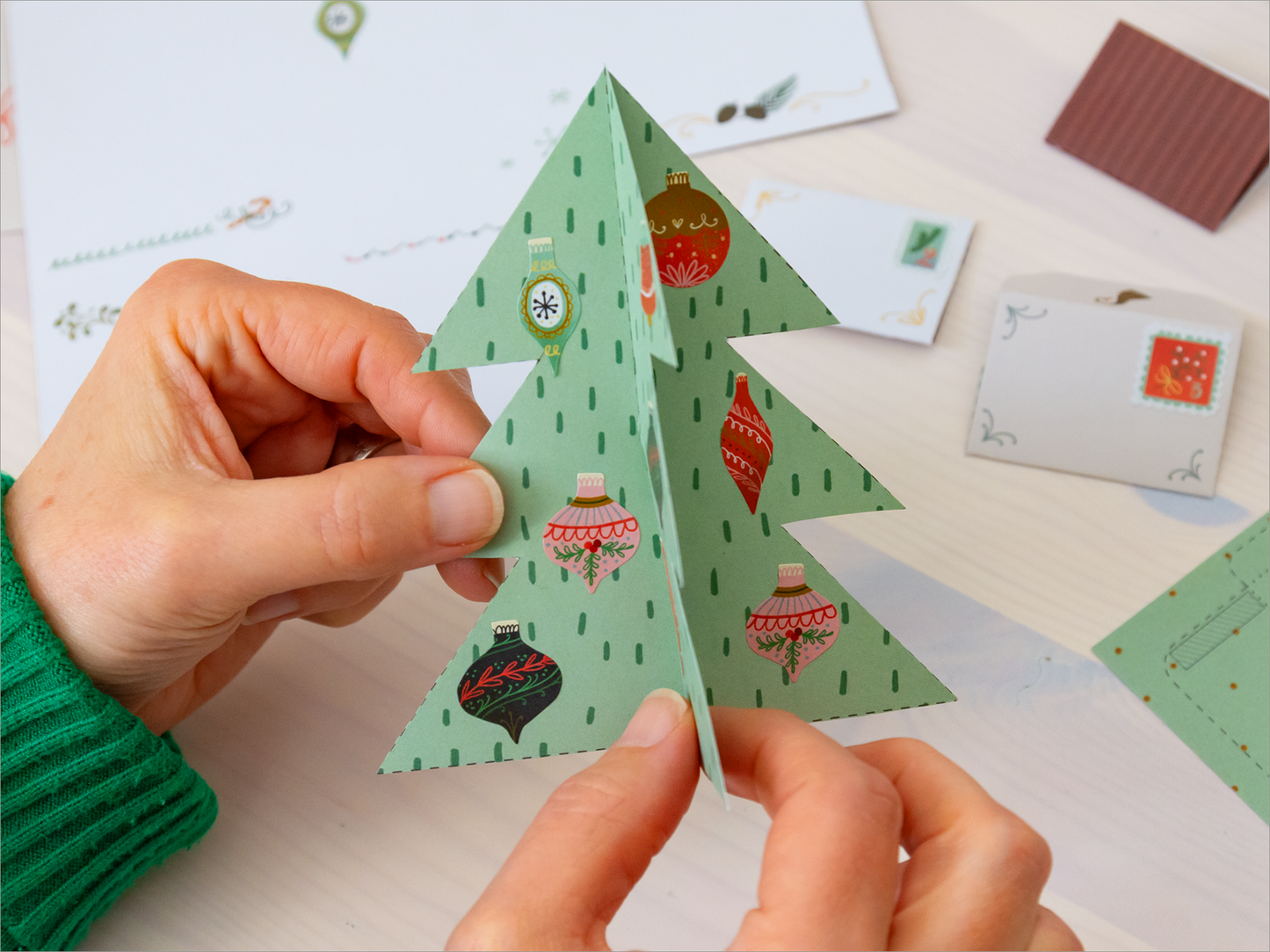 Die Innenseite des Buches „Der Wichtelblock“ zeigt eine zwei Kinderhände, die einen handgefertigten, dreidimensionalen Weihnachtsbaum aus grünem Papier mit bunten Kugeln halten. Im Hintergrund sind weitere Bastelmaterialien sichtbar, darunter kleine Kärtchen und Dekorationen. Die Farben sind hauptsächlich grün, rot und gold, die eine festliche Atmosphäre schaffen.