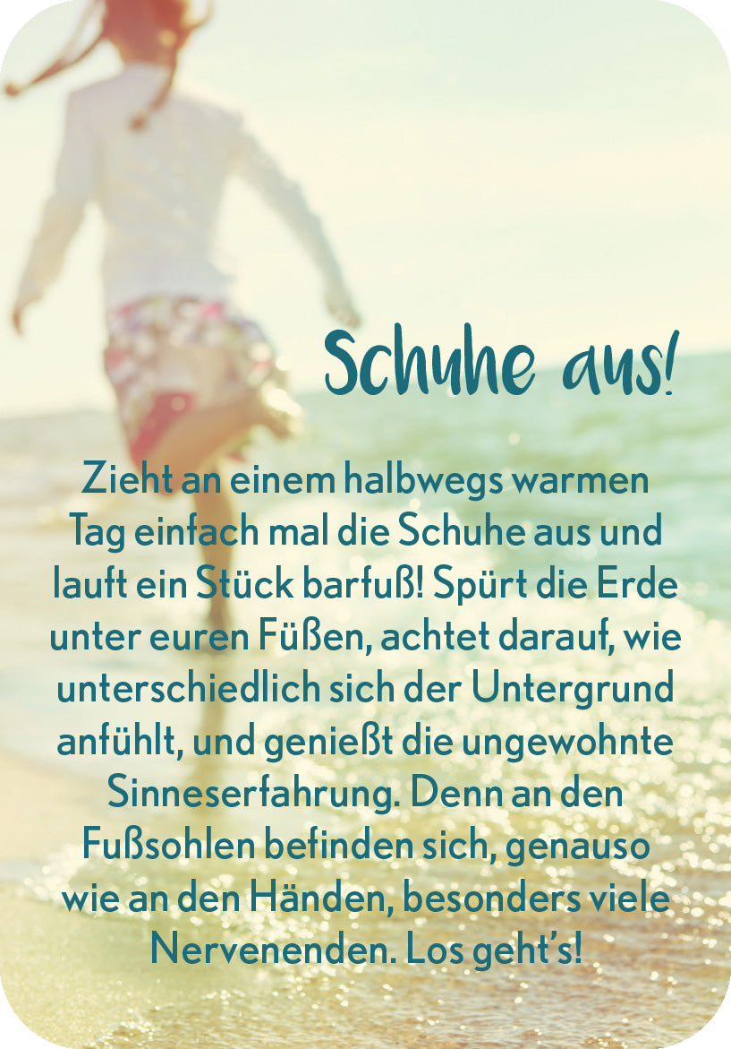 Gezeigt wird eine Innen- oder Zusatzansicht von „Achtsamkeit in der Familie“. Die Gestaltung zeigt einen verschwommenen Hintergrund am Strand mit bläulichen und sandfarbenen Tönen, die eine entspannte Atmosphäre schaffen. Im Vordergrund steht der handschriftlich gestaltete Text „Schuhe aus!“, der in einer verspielten, grafischen Schriftart in Blau gehalten ist. Darunter folgt in einer klaren, gut lesbaren Serifenschrift der Anleitungstext in Schwarz, der zur Sinneserfahrung einlädt.