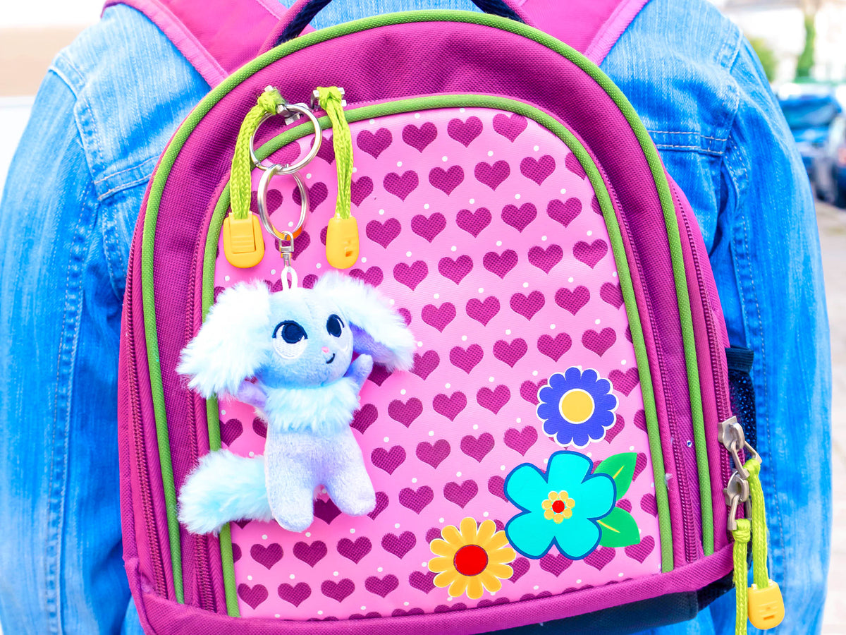 Gezeigt wird eine Innen- oder Zusatzansicht von „Ein Inne halten“. Der Rucksack zeigt ein lebhaftes, pinkfarbenes Design mit Herzen und farbenfrohen Blumenmotiven. Im Vordergrund hängt ein flauschiger Schlüsselanhänger in Form eines lilafarbenen Tierchens. Die Reißverschlusszieher sind in leuchtendem Gelb gehalten. Die Farbgebung und die verspielte Typografie vermitteln eine fröhliche, kindgerechte Atmosphäre.