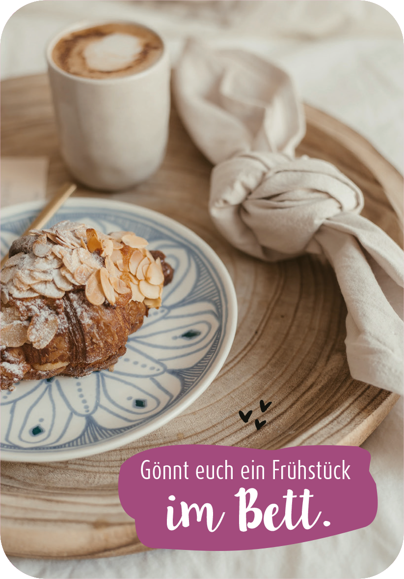 Gezeigt wird eine Innen- oder Zusatzansicht von „99 herrlich verrückte Ideen für euer Leben zu zweit“. Die Darstellung zeigt ein rustikales Holztablett mit einem Croissant, das mit Mandelblättchen garniert ist, und einer Tasse Kaffee. Der Hintergrund ist hell und neutral gehalten. Der Text „Gönnt euch ein Frühstück im Bett.“ ist in einer verspielten, weißen Schrift auf einem lila Hintergrund platziert, ergänzt durch kleine Herzsymbole.