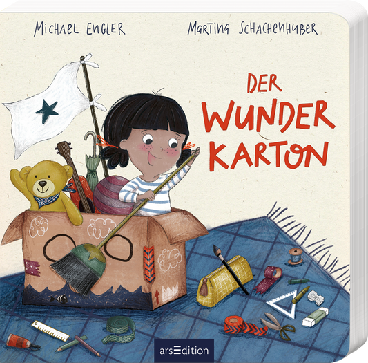 Das Buchcover zeigt ein liebevoll illustriertes Mädchen mit schwarzem Haar, das freudig aus einem großen, offenen Karton blickt. Der Karton ist gefüllt mit verschiedenen bunten Materialien, darunter ein Teddybär, eine Pinsel und andere Bastelutensilien. Über dem Mädchen weht eine kleine weiße Flagge mit einem schwarzen Stern. Die Schriftzüge „DER WUNDER KARTON“ sind in auffälligem Rot gestaltet. Oben stehen die Namen der Autoren: Michael Engler und Marting Schachenhuber. Ganz unten...