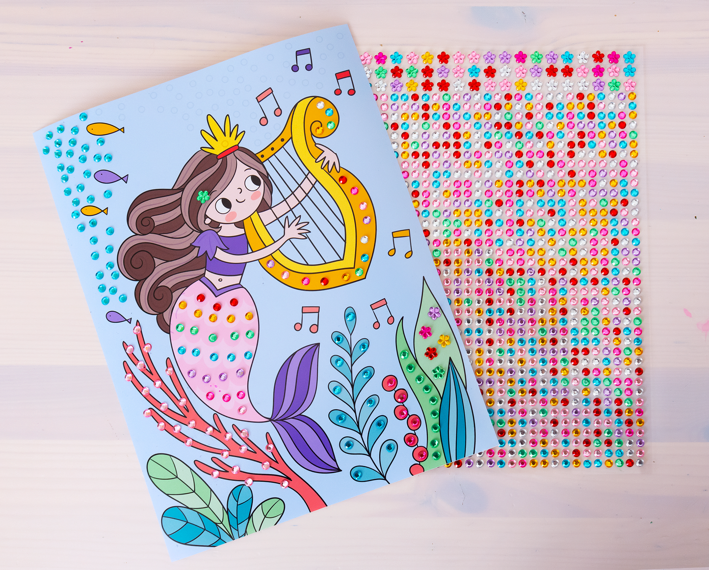 Gezeigt wird eine Innen- oder Zusatzansicht von „Mein Diamant-Stickerheft – Meerjungfrauen“. Zu sehen sind zwei herausgetrennte Seiten, die auf einem weißen Holztisch liegen. Die Seite mit der Szene zeigt eine fröhliche Meerjungfrau mit braunen Haaren, die eine goldene Harfe spielt. Umgeben ist sie von bunten, stilisierten Fischen und Wasserpflanzen. Die Farbpalette umfasst sanfte Blautöne, lebhafte Pink- und Lilanuancen sowie leuchtendes Gelb. Darunter hervor lugt ein Stickerbogen mit...