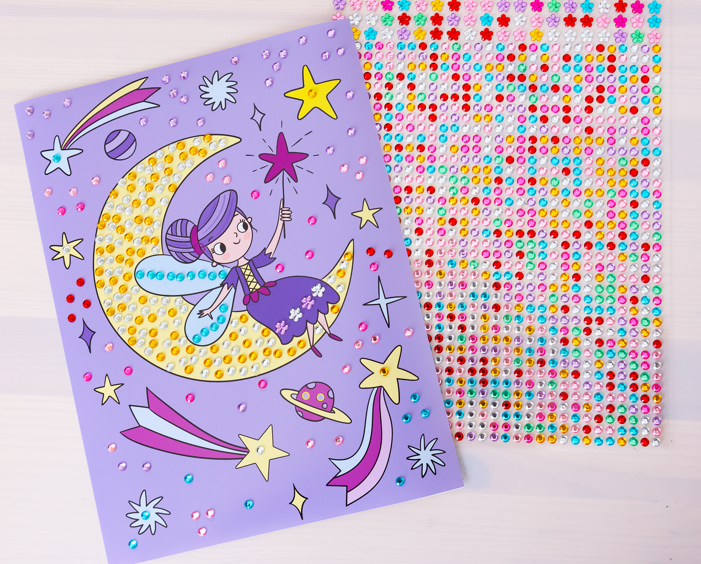 Gezeigt wird eine Innen- oder Zusatzansicht von „Mein Diamant-Stickerheft – Feen“. Es werden zwei herausgetrennte Seiten gezeigt. Das obere Blatt zeigt eine lilafarbene Seite mit einer fröhlichen Fee, die auf einem Mond sitzt und einen Zauberstab hält. Umgeben ist sie von bunten, glitzernden Stickern sowie Sternen und Planeten. Die Farben sind lebhaft, mit vielen Pastelltönen und glitzernden Akzenten. Der Stickerbogen lugt darunter hervor und zeigt die vielen bunten Diamantsticker.