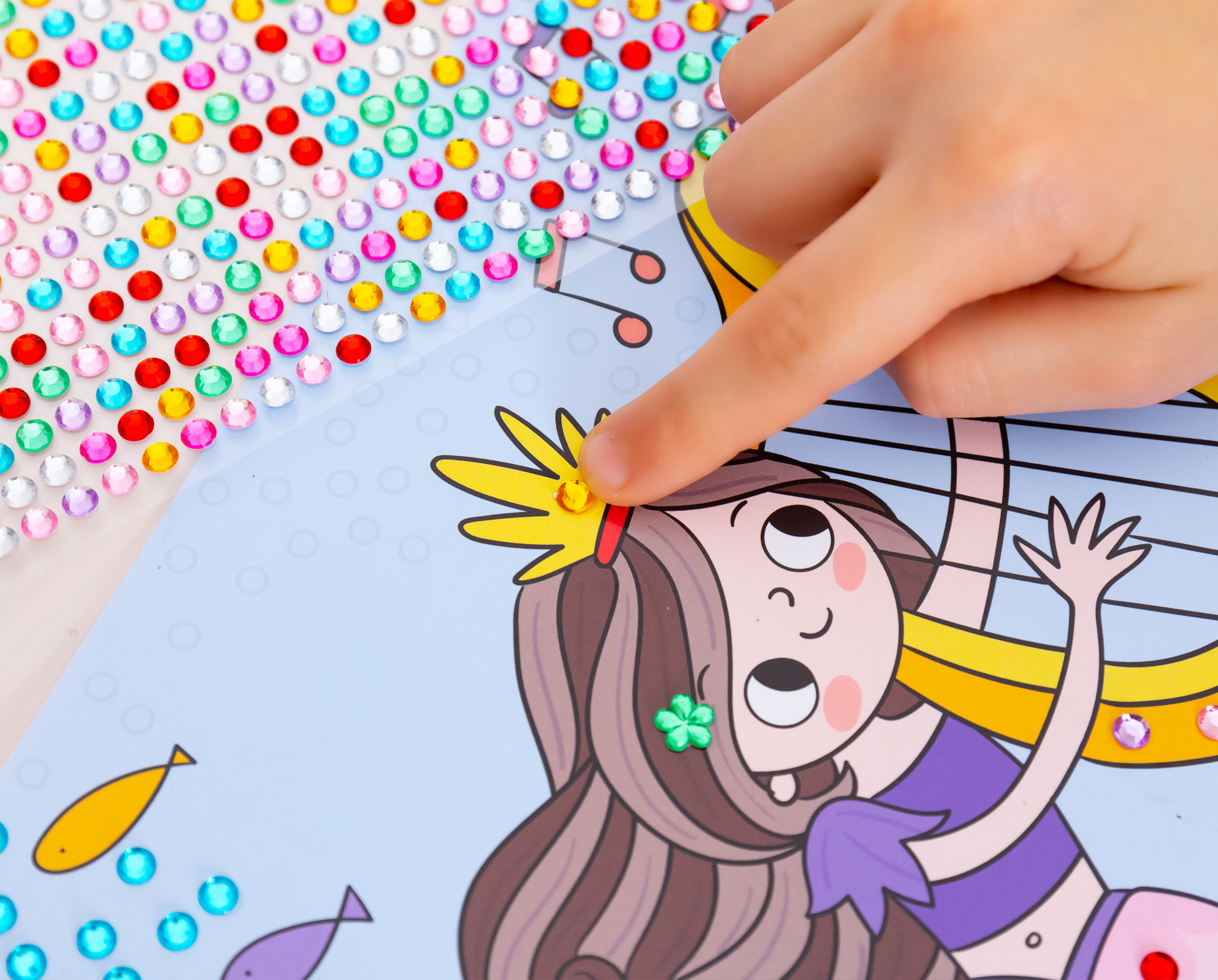 Gezeigt wird eine Innen- oder Zusatzansicht von „Mein Diamant-Stickerheft – Feen“. Aus der oberen rechten Ecke ragt eine Kinderhand in das Bild, welche einen gelben Diamantsticker auf der Krone einer Meerjungfrau platziert. In der oberen linken Ecke sieht man einen Teil des Stickerbogens auf dem eine Vielzahl von bunten Diamantstickern zur Verfügung stehen.