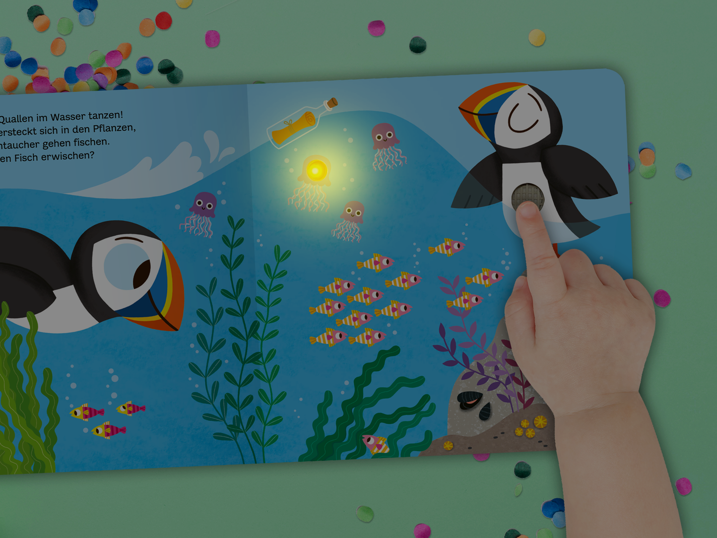 Gezeigt wird eine Innen- oder Zusatzansicht von „Mein blinkendes Soundbuch – Am Meer“. Die Seite zeigt eine farbenfrohe Unterwasserwelt mit zwei fröhlichen Papageitauchern, die in hellen Blau- und Grüntönen abgebildet sind. Bunte Fische schwimmen entlang einer stilisierten Pflanzenwelt. Ein leuchtendes Icon zeigt, wo die Klänge aktiviert werden können. Die spielerische Typografie ermuntert zum Mitmachen und Entdecken.