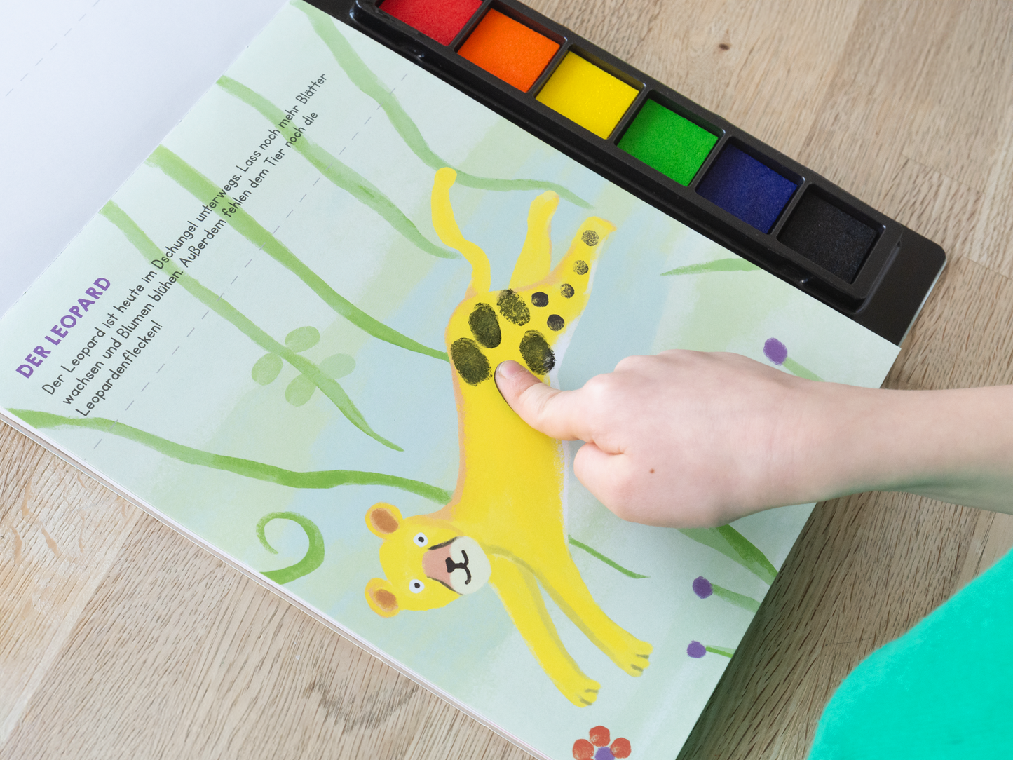 Gezeigt wird eine Innen- oder Zusatzansicht von „Meine allerersten Fingerstempel Tiere“. Auf der Seite ist ein gelber Leopard abgebildet, der mit grünen Sträuchern und bunten Blumen in einem hellen, freundlichen Umfeld platziert ist. Die Typografie ist klar und gut lesbar, mit dem Titel „DER LEOPARD“ in einer kindgerechten Schriftart. Ein Kinderfinger ist gerade dabei, Farbe auf den Leoparden zu stempeln, was eine interaktive Element verdeutlicht.