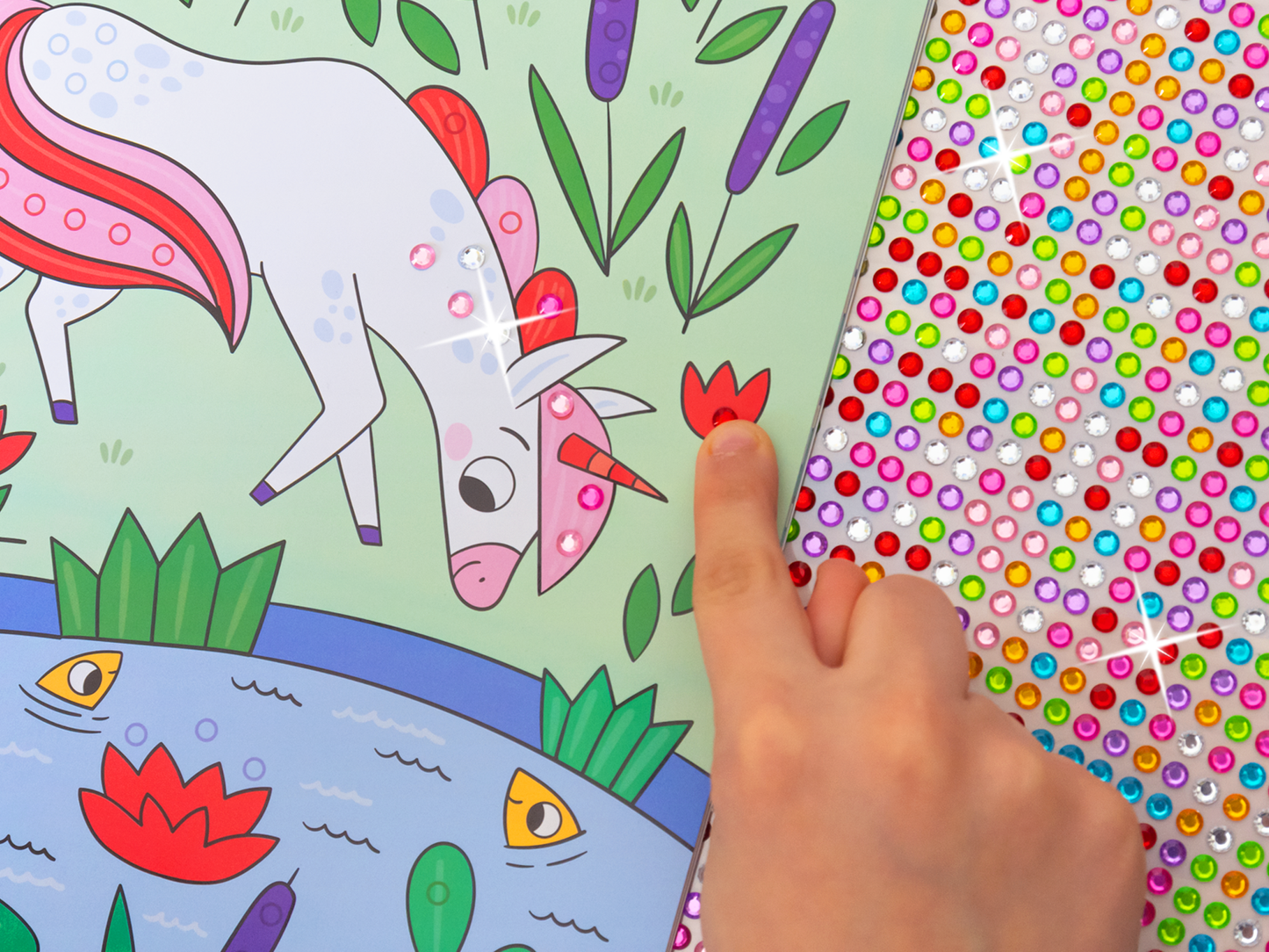 Es wird eine Innenseite von „Mein Diamant-Stickerheft – Einhörner“ gezeigt, wie es von einer Kinderhand bestickert wird. Der Diamantstickerbogen liegt rechts neben der Innenseite unter zeigt eine große Vilezahl an Farben. Das Kind klebt soeben einen roten Sticker auf eine Blume. Weiter ist auf dem Bild ein Einhorn zu sehen, das auf einer grünen Wiese mit lila Blumen und einem blauen Teich steht, in dem sich Seerosen befinden.