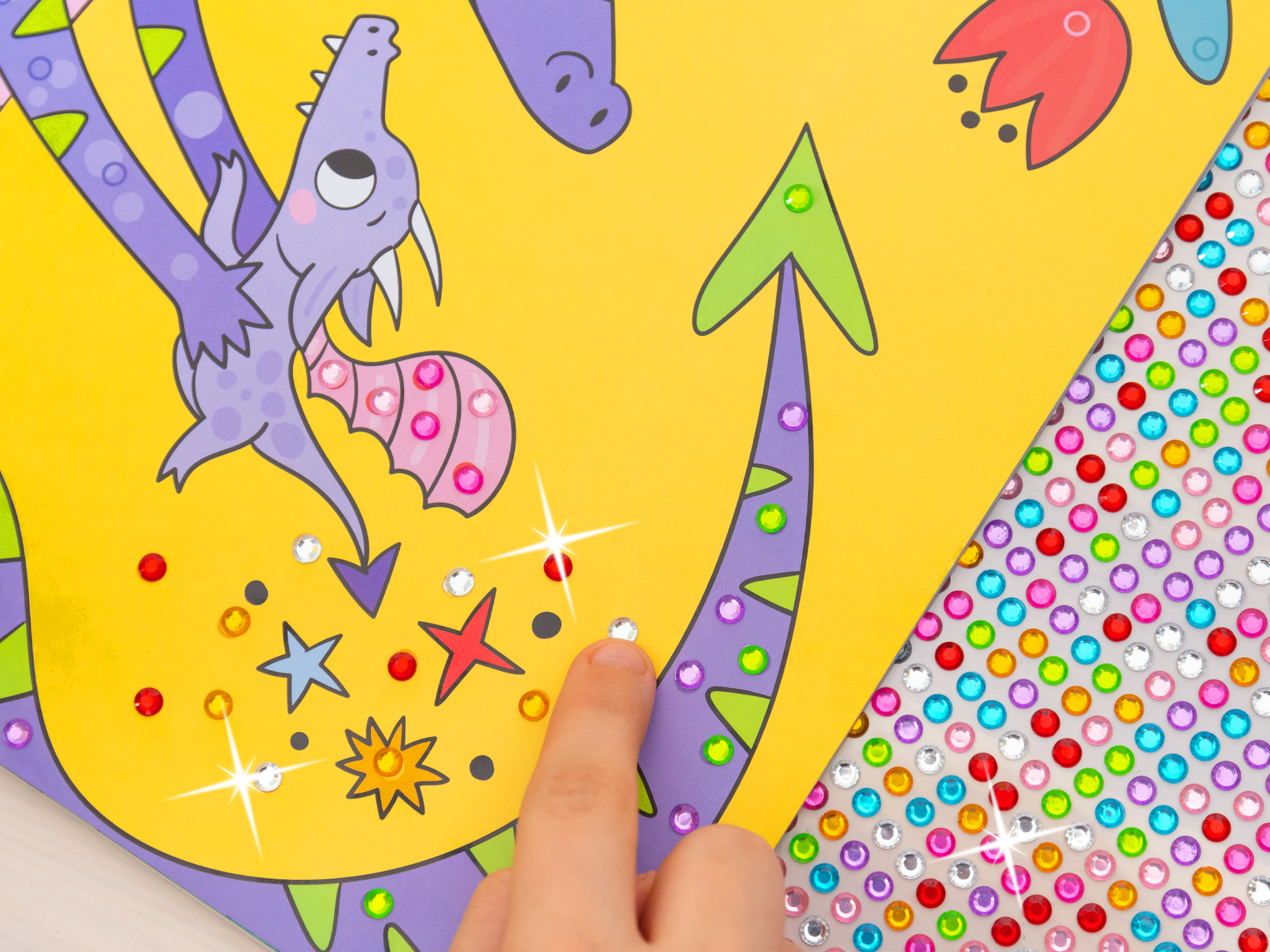 Es wird eine Innenseite von „Mein Diamant-Stickerheft – Drachen“ in Aktion gezeigt. Eine Kinderhand klebt soeben einen weißen Diamantsticker auf eine Innenseite des Heftes. Rechts daneben liegt der Stickerbogen mit einer großen Vielfalt an farbigen Steinchen.