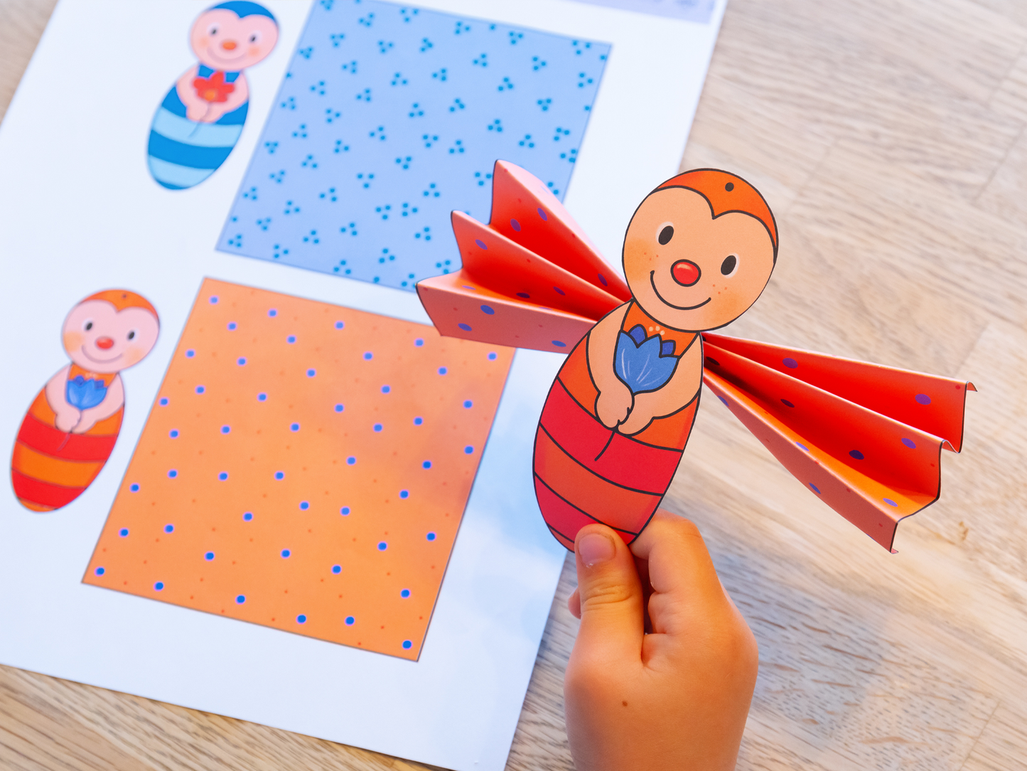 Gezeigt wird eine Innen- oder Zusatzansicht von „Mein erstes großes Ferien-Bastelheft“. Im Vordergrund wird eine bunte, selbstgemachte Figur in Form eines Insekts präsentiert, die in warmen Farben wie Rot und Orange gestaltet ist. Im Hintergrund sind zwei Papierblätter mit unterschiedlichen Mustern: eines mit blauen Punkten auf leichtem Hintergrund und das andere in Orange mit blauen Punkten. Die fröhliche Typografie unterstreicht die kinderfreundliche Atmosphäre des Hefts.