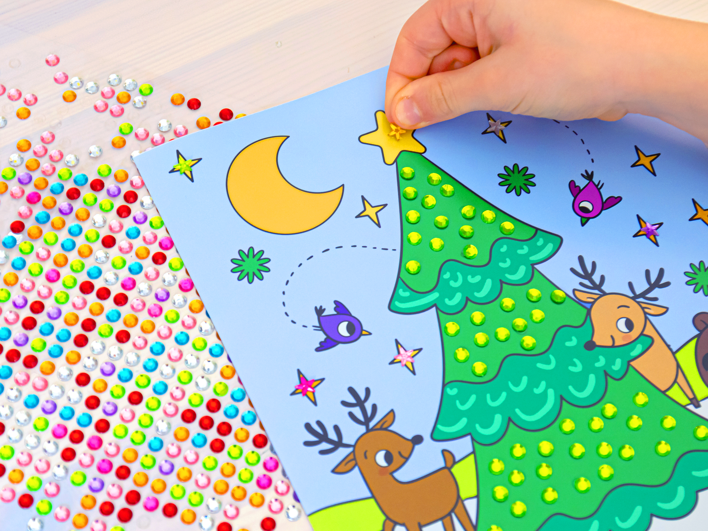 Gezeigt wird eine Innen- oder Zusatzansicht von „Mein Diamant-Stickerheft – Wintertraum“. Eine Innenseite wird hier in Aktion gezeigt, denn eine Kinderhand ist soeben dabei, einen Sticker aufzukleben. Alle anderen Felder sind bereits mit grünen Diamantstickern beklebt und bringen das Bild zu glitzern und funkeln. Links neben der Seite sieht man den Stickerbogen, auf dem eine bunte Vielzahl an Diamantstickern zu erkennen ist.