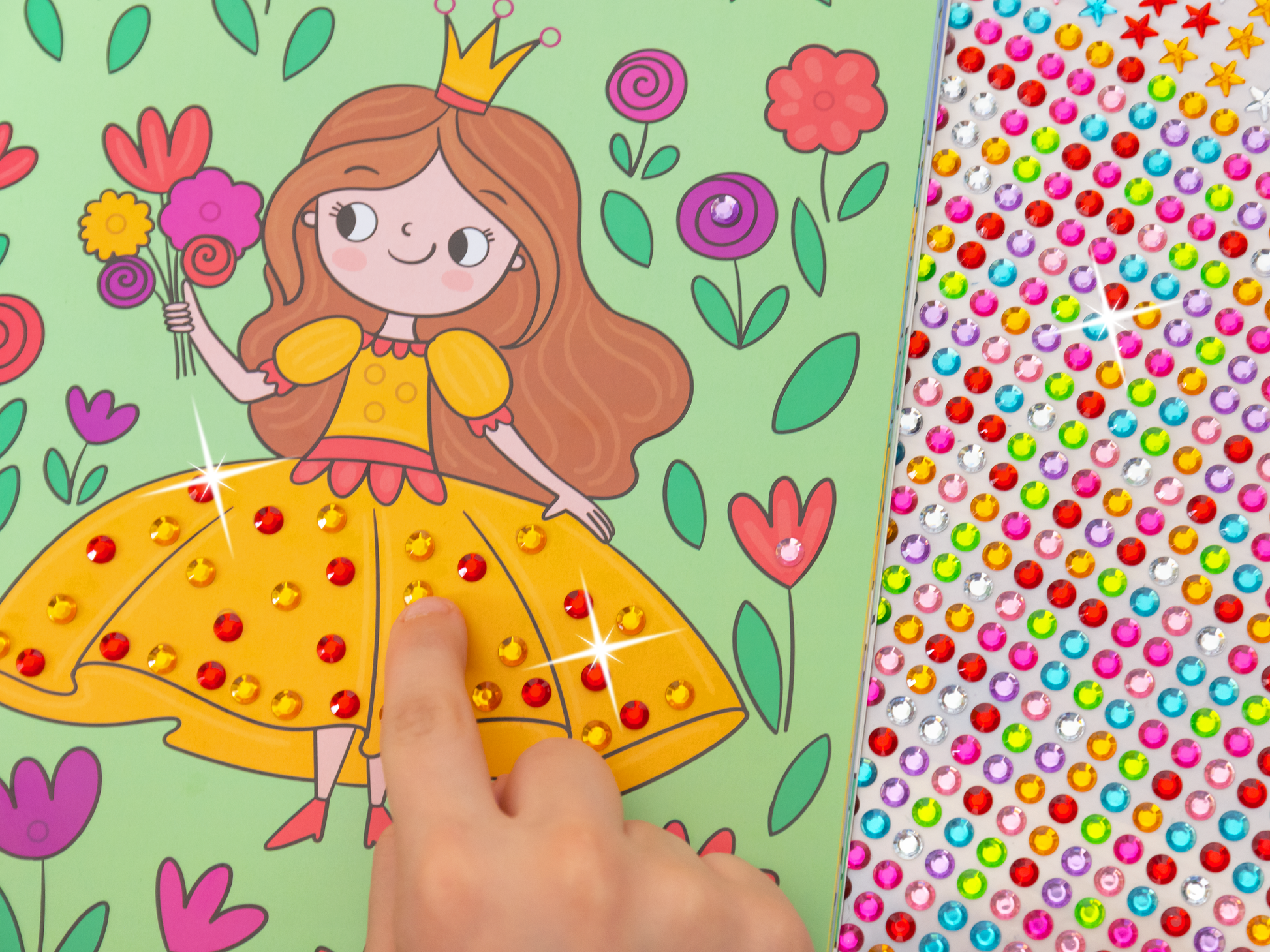 Auf der Innenseite des Buches „Mein Diamant-Stickerheft – Prinzessinnen“ ist eine fröhliche Prinzessin mit langen, braunen Locken zu sehen, die ein leuchtend gelbes Kleid trägt. Ihre Krone funkelt und sie hält einen Blumenstrauß in der Hand. Um sie herum sind bunte Blumen in verschiedenen Farbtönen. Rechts ist eine Vielzahl von Glitzerstickern in verschiedenen Farben und Formen angeordnet, die die Kreativität anregen. Eine Kinderhand klebt gerade einen Diamanten auf das Bild.