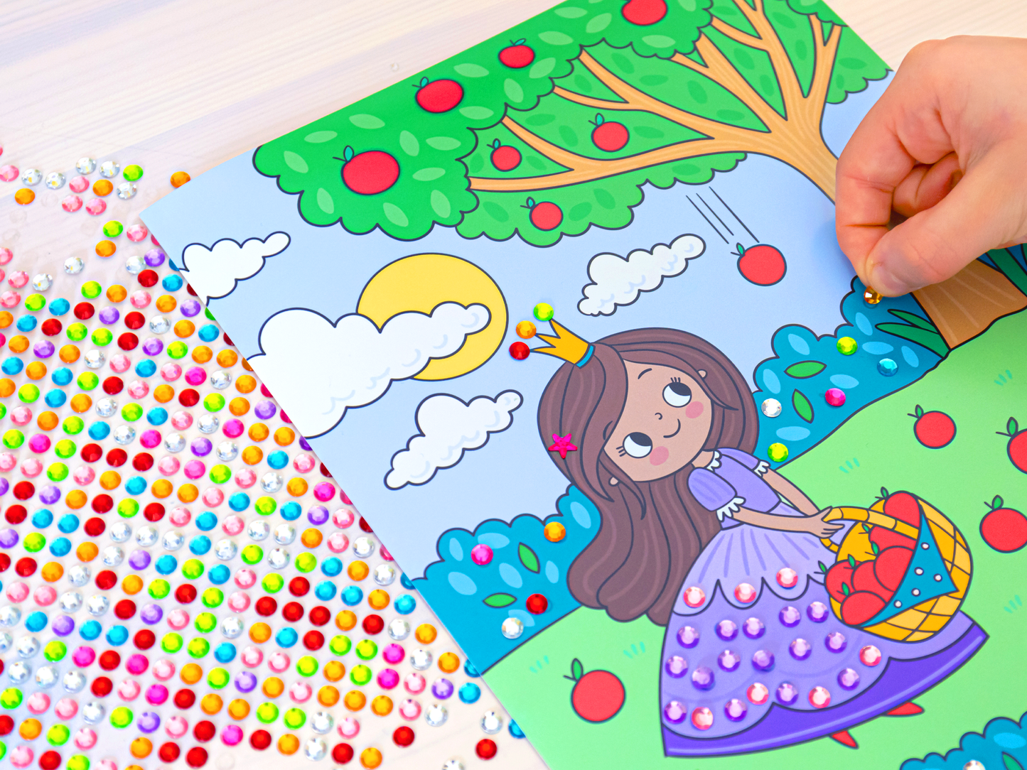 Gezeigt wird eine Innen- oder Zusatzansicht von „Mein Diamant-Stickerheft – Prinzessinnen“. Die Abbildung zeigt eine fröhliche Prinzessin in einem lila Kleid, die einen Korb mit Äpfeln hält. Umgeben von einem bunten Hintergrund mit grünen Bäumen, roten Äpfeln und ein paar Wolken unter einem leuchtenden, gelben Sonne. Auf der linken Seite sind zahlreiche glitzernde Sticker in verschiedenen Farben verteilt, die zum Bekleben einladen. Die Hand eines Kindes klebt soeben einen...