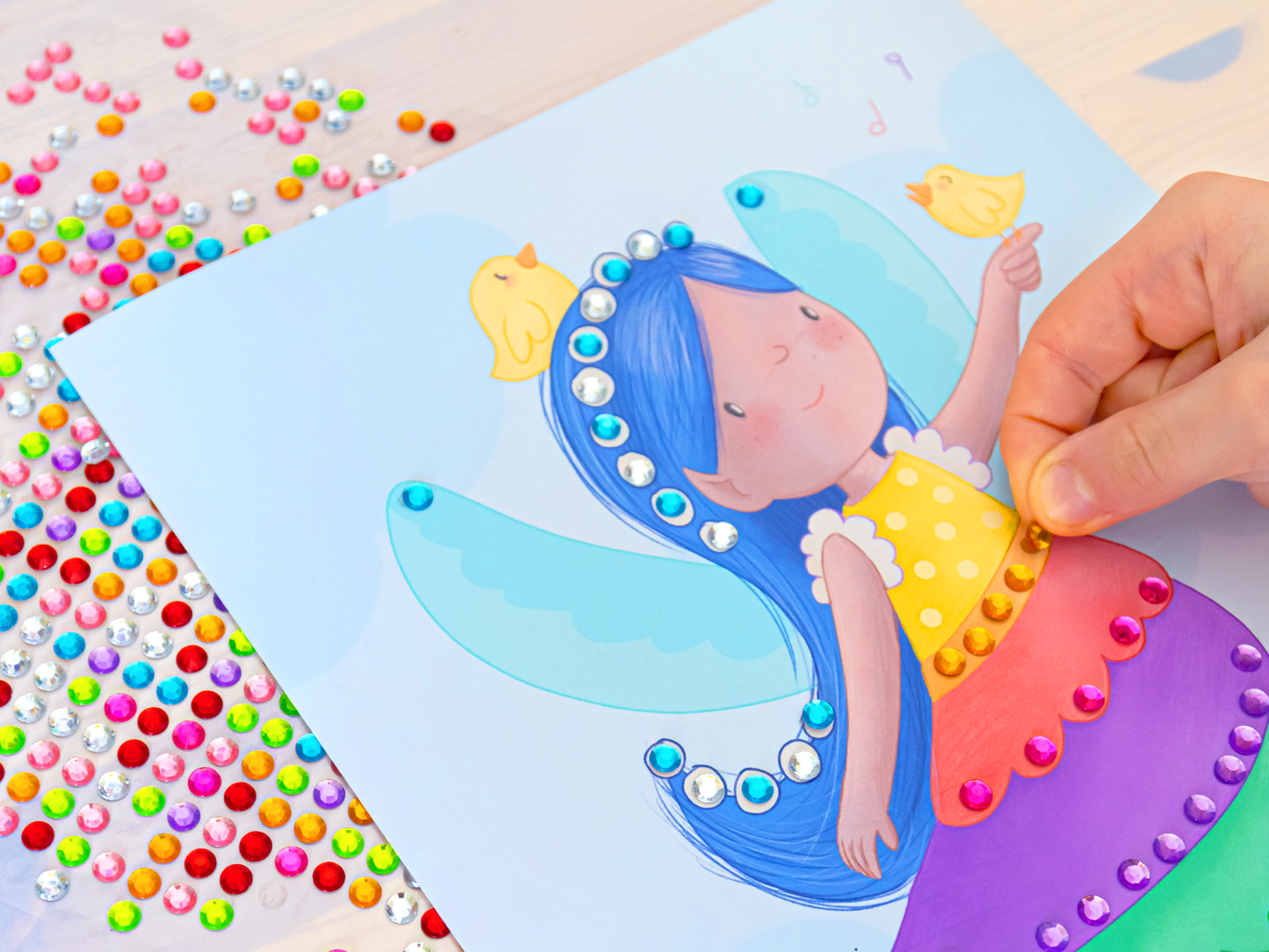 Gezeigt wird eine Innen- oder Zusatzansicht von „Diamantsticker Anziehpuppen – Feenzauber“. Das Bild zeigt eine farbenfrohe Darstellung einer Feenfigur mit blauen Flügeln und langen blauen Haaren. Sie trägt ein gelbes Oberteil mit weißen Rüschendetails. Um die Figur herum sind bunte, funkelnde Diamantsticker in verschiedenen Farben verteilt. Eine Hand platziert gerade einen Sticker auf das Bild. Der Hintergrund ist hellblau und verleiht der Szene eine fröhliche Atmosphäre. Unter der...