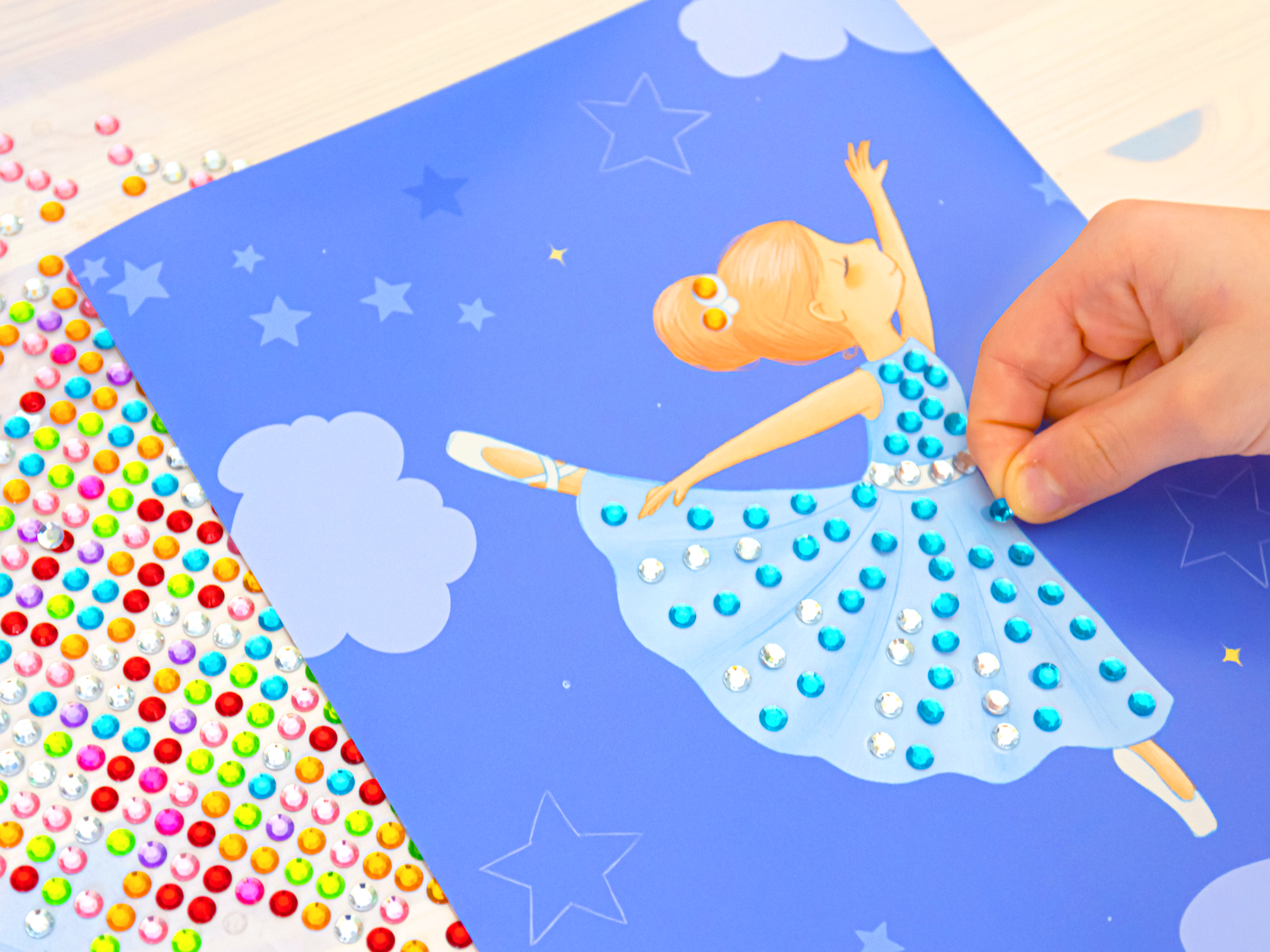 Gezeigt wird eine Innen- oder Zusatzansicht von „Diamantsticker Anziehpuppen – Ballett“. Im Bild ist eine ballerinaartige Figur auf einem hellblauen Hintergrund mit Wolken und Sternen dargestellt. Die Ballerina trägt ein schwingendes Kleid mit blauen und weißen Glitzersteinen, die von einem Kind gesetzt wurden, dessen Hand man im Bild sieht. Neben der Figur befinden sich farbenfrohe Aufkleber in verschiedenen Größen, die zur Gestaltung verwendet werden können.