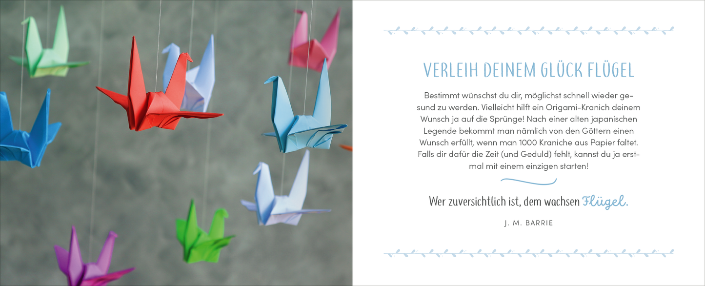 Gezeigt wird eine Innen- oder Zusatzansicht von „Gute Besserung – Alles Liebe!“. Auf der linken Seite hängen bunt gefaltete Papierkraniche in verschiedenen Farben wie Rot, Blau, Grün und Pink vor einem grauen Hintergrund. Die rechte Seite ziert ein eleganter Text in Türkis mit der Überschrift „Verleih deinem Glück Flügel“ sowie einem Zitat von J. M. Barrie. Die Schrift ist klar und modern, um eine freundliche und optimistische Stimmung zu vermitteln.
