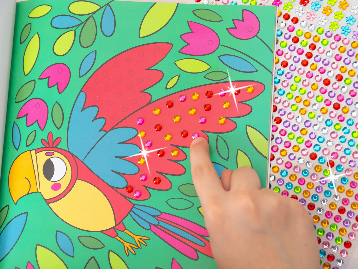 Gezeigt wird eine Innen- oder Zusatzansicht von „Mein Diamant-Stickerheft – Tiere“. Zu sehen ist einen Innenseite in Aktion, denn eine Kinderhand klebt soeben einen Diamantsticker auf. Neben dem Heft liegt der Stickerbogen mit der bunten Vielzahl an Glitzersteinen. Die Innenseite zeigt einen bunten Papageien mit leuchtenden roten und blauen Federn auf einem lebhaften, grünen Hintergrund, umgeben von bunten Blüten.