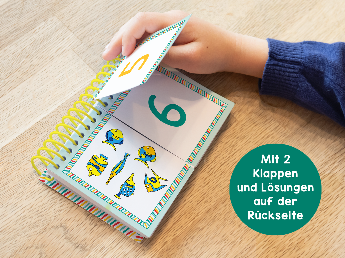 Gezeigt wird eine Innen- oder Zusatzansicht von „Lernraupe – Mix-Max Erste Zahlen“. Die Darstellung zeigt den offenen Lernblock mit einem großen, grünen Ziffern „6“ auf der linken Seite. Darunter befinden sich bunte Illustrationen von Fischen und anderen Tieren. Eine Hand hält eine Klappenkarte mit der Ziffer „2“ darüber. Der Hintergrund des Blocks ist hell und der Rand ist mit bunten, gestreiften Mustern verziert. In einem grünen Kreis steht: „Mit 2 Klappen und Lösungen...