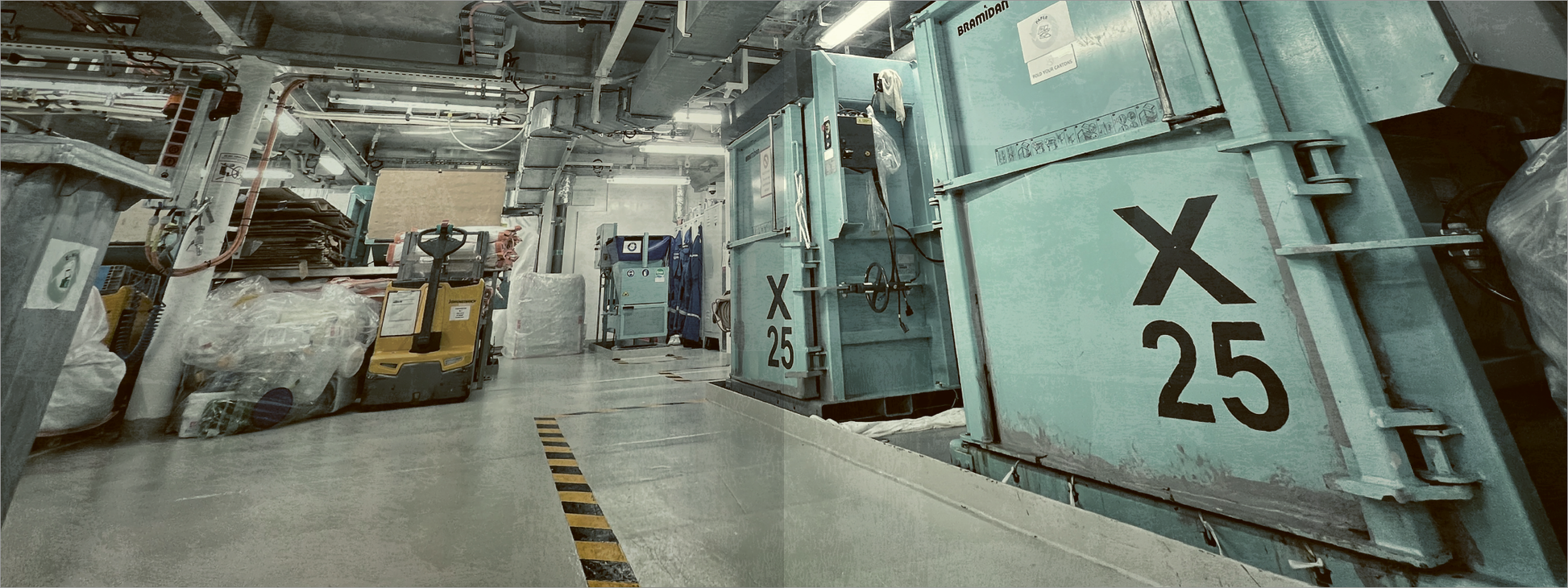 Gezeigt wird eine Innen- oder Zusatzansicht von „Escape Room. Christmas Cruise – Kreuzfahrt in den Tod“. Die Szene präsentiert einen industriell gestalteten Raum, dominiert von großen, blaugrünen Abfallcontainern mit der Aufschrift „X 25“. Der Boden ist glatt und grau, während gelbe Sicherheitsmarkierungen den Weg anzeigen. Die Beleuchtung ist kühl und sorgt für eine sterile Atmosphäre. Im Hintergrund sind weitere Lagereinheiten und ein Gabelstapler sichtbar, die die...