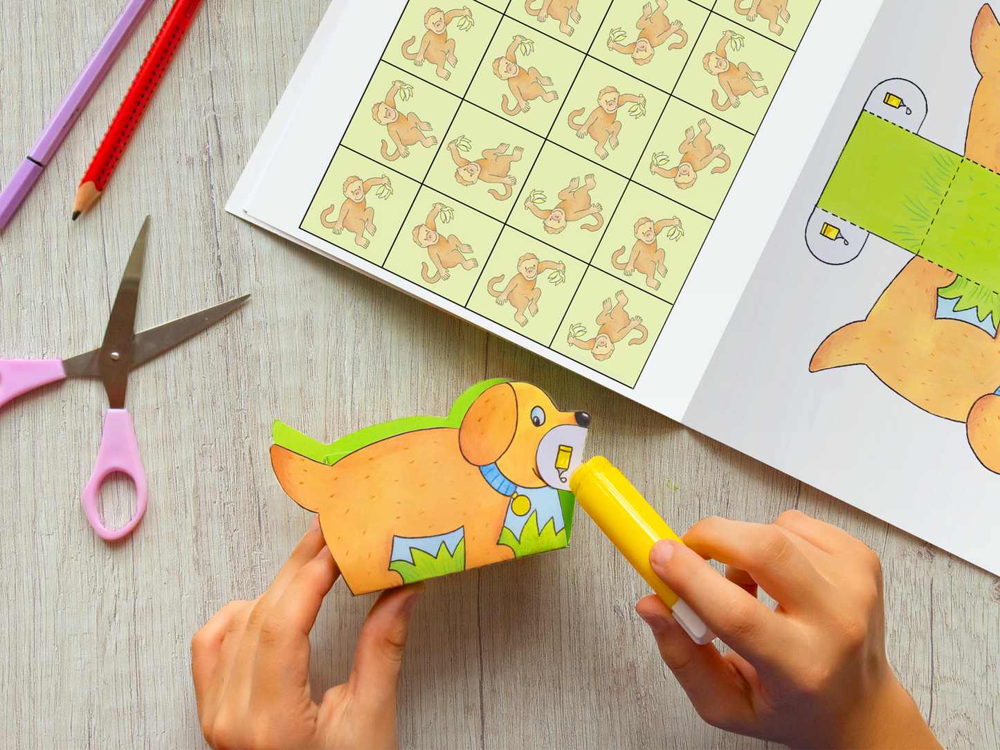 Gezeigt wird eine Innen- oder Zusatzansicht von „Mein erstes großes Tier-Bastelheft“. Im Vordergrund sind die Hände eines Kindes zu sehen, die eine mit Farben gestaltete Hundefigur halten. Die Figur ist in Hellbraun, mit grünen Akzenten auf dem Rücken, orange Punkten und einem blauen Halstuch. Im Hintergrund befindet sich ein leicht geöffneter Bastelblock mit farbenfrohen Tierillustrationen und Schnittmustern, umgeben von bunten Buntstiften und einer Schere. Die Typografie ist...