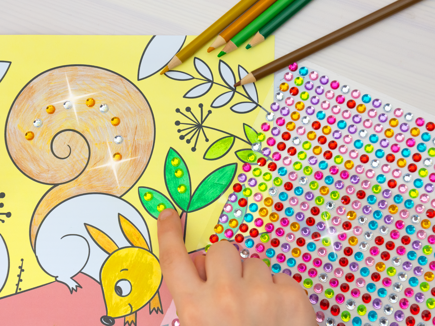 Gezeigt wird eine Innen- oder Zusatzansicht von „Diamant-Malbuch Tiere“. Die Abbildung zeigt eine Innenseite, die gerade von einem Kind bestickert und angemalt wird. Ein niedliches Eichhörnchen und stilisierte Pflanzen sind klar erkennbar. Bunte Stifte ragen von oben in das Bild. Glitzernde Strasssteine liegen auf einem Tisch und ergänzen das Malbild. Eine Kinderhand klebt einen grünen Diamanten auf ein Blatt.