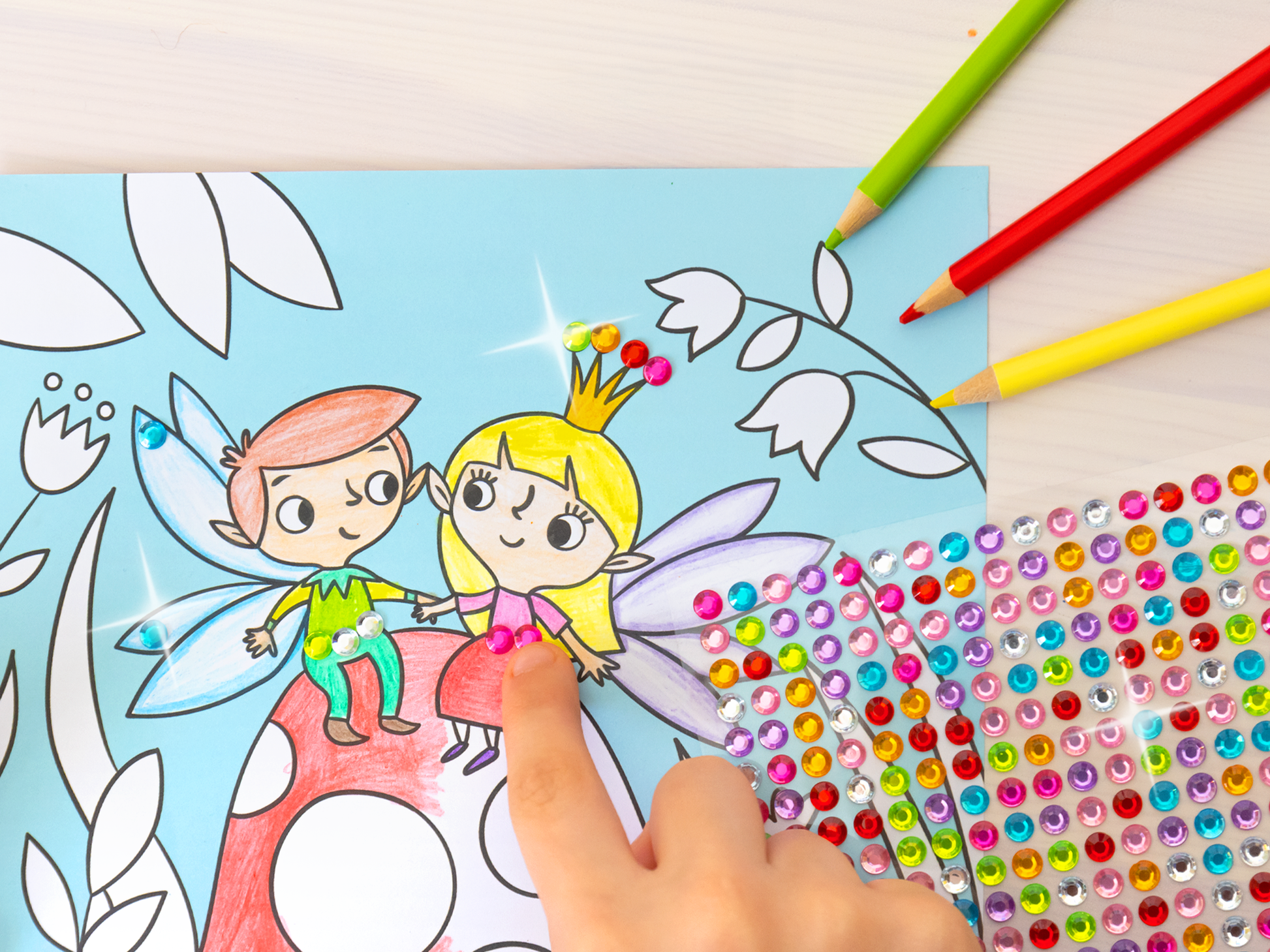 Gezeigt wird eine Innen- oder Zusatzansicht von „Diamant-Malbuch Feen“. Die Illustration zeigt zwei fröhliche Feen auf einem großen, roten Fliegenpilz, umgeben von grünen Blättern und bunten Details. Eine Kinderhand ragt von unten in das Bild und stickert gerade einen Diamanten auf das Bild. Von rechts ragen Buntstifte und ein Stickerbogen in das Bild.