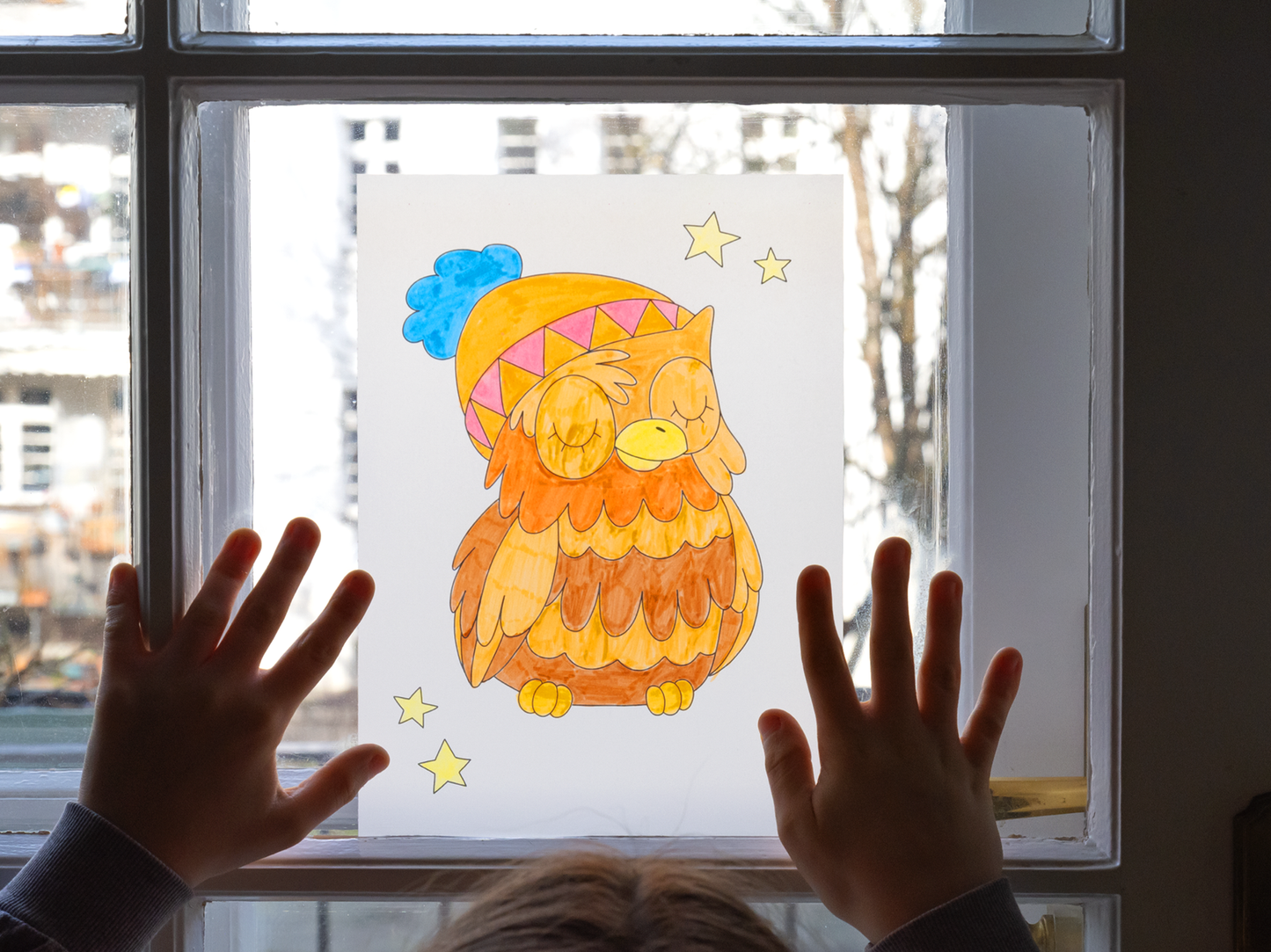 Gezeigt wird eine Innen- oder Zusatzansicht von „Ausmal-Fensterbilder – Im Winter“. Die Darstellung zeigt eine bunte Eule mit einer gestrickten Mütze in Blau und Orange, umrahmt von gelben Sternen. Die Eule hat geschlossene Augen und ein freundliches Aussehen. Das Bild wird von einem Kind an eine Glasscheibe gedrückt. Die Farben sind leuchtend und warm, was eine gemütliche Winterstimmung vermittelt. Der Hintergrund zeigt unscharf die Außenwelt.