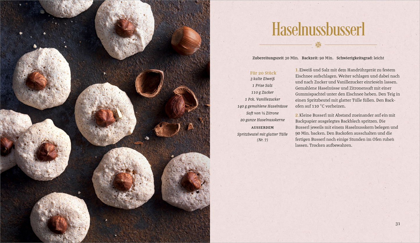 Gezeigt wird eine Innen- oder Zusatzansicht von „Vanille, Zimt und Weihnachtsduft“. Auf der linken Seite sind mehrere runde, helle Haselnussplätzchen zu sehen, die jeweils mit einer ganzen Haselnuss darauf dekoriert sind. Der Hintergrund ist dunkel und strukturiert, was einen ansprechenden Kontrast zu den Plätzchen bildet. Rechts befindet sich ein Rezepttext in eleganter, goldfarbener Schrift, der auf einem sanft rosa Hintergrund steht und eine leicht lesbare, moderne Typografie verwendet.