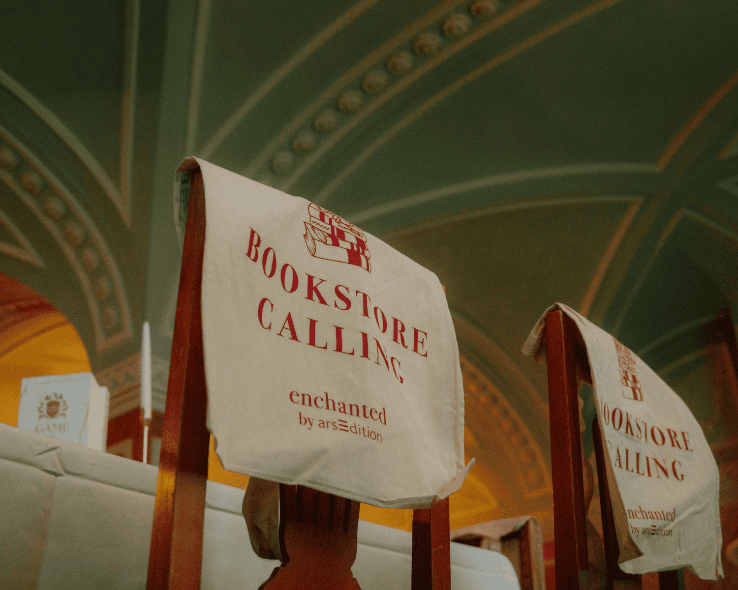 Fotovon Stuhllehnen über denen Jutebeutel mit der Aufschrift "Bookstore calling" hängen. Im Hintergrund Gewölbedecken.