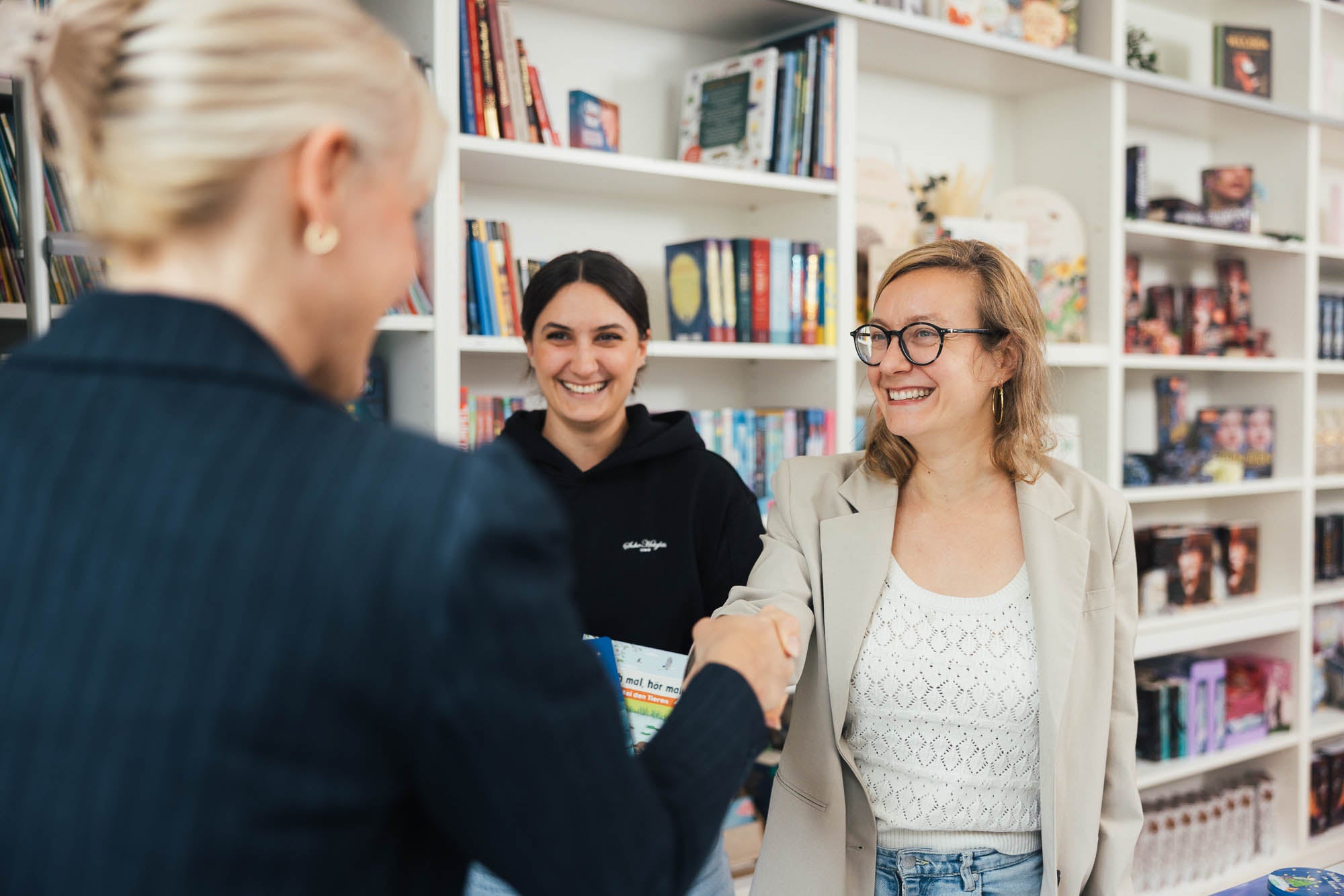 Zwei Personen schütteln sich in einem hellen Raum mit Bücherregalen die Hand, symbolisch für eine geschäftliche Vereinbarung im Lizenzbereich. Im Hintergrund steht eine weitere Person und verfolgt das Gespräch. Die Regale sind mit bunten Büchern gefüllt und schaffen eine professionelle, aber einladende Atmosphäre.