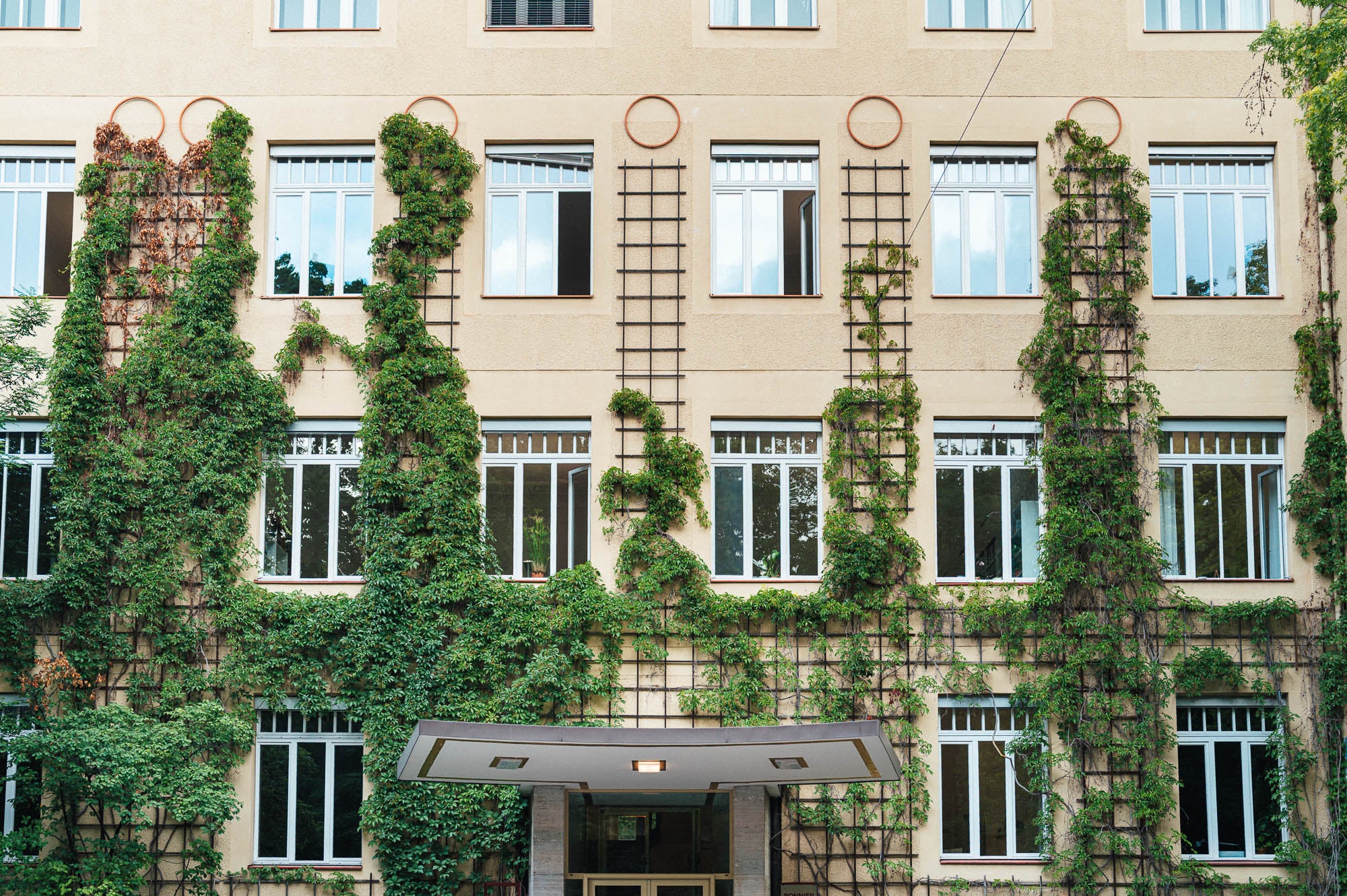Das Verlagsgebäude der arsEdition mit einer beigefarbenen Fassade, die durch rankendes Grün an Metallgittern begrünt ist. Gleichmäßig angeordnete Fenster prägen die Fassade, einige davon sind geöffnet. Über dem Eingang befindet sich ein modernes Vordach mit integrierter Beleuchtung. Die Begrünung verleiht dem Gebäude eine einladende und nachhaltige Ausstrahlung.