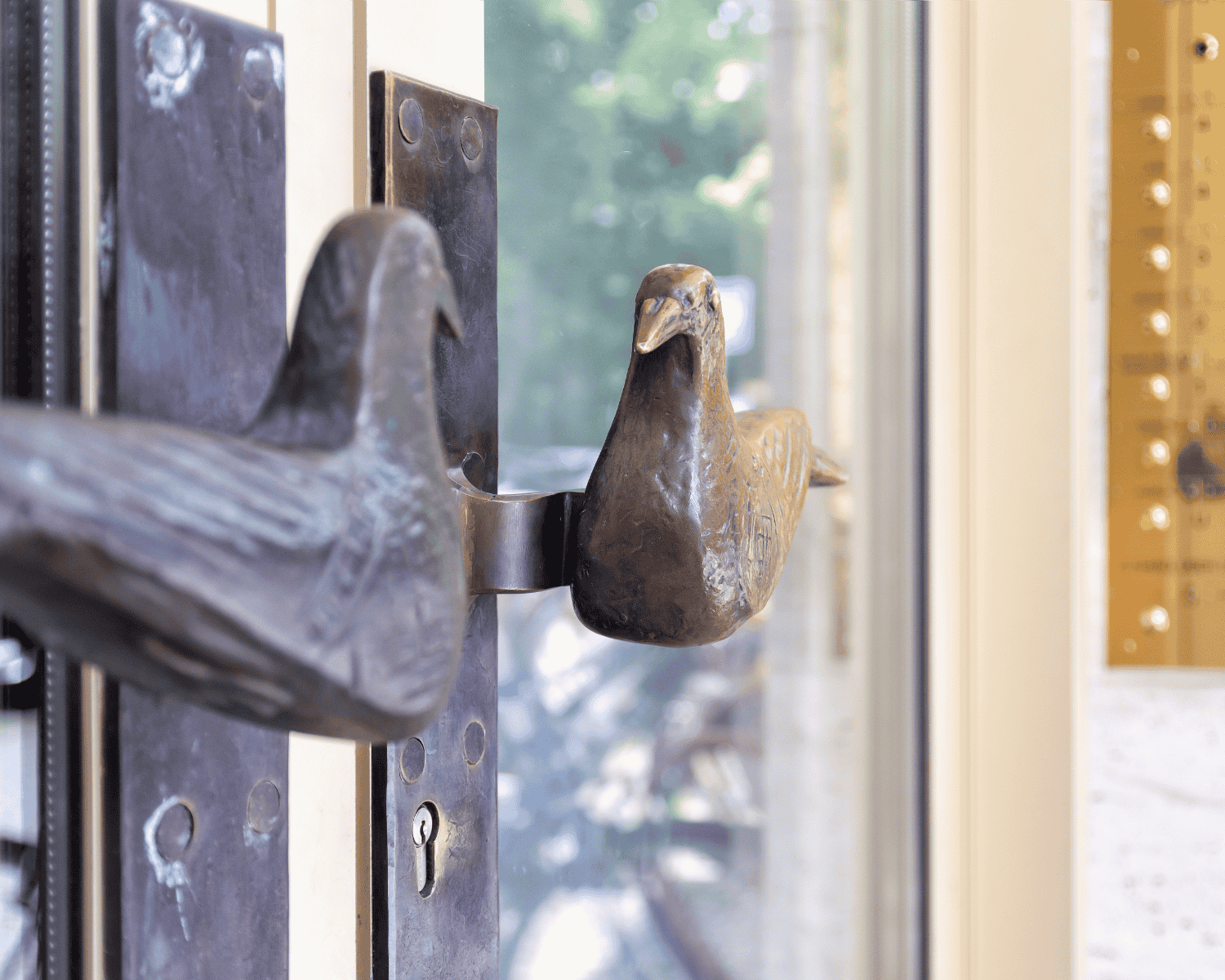 Detailaufnahme eines Türgriffs in Form eines Vogels aus Metall, befestigt an einer Glastür. Im Hintergrund sind verschwommene grüne Bäume und ein Teil eines goldfarbenen Türschilds zu sehen.