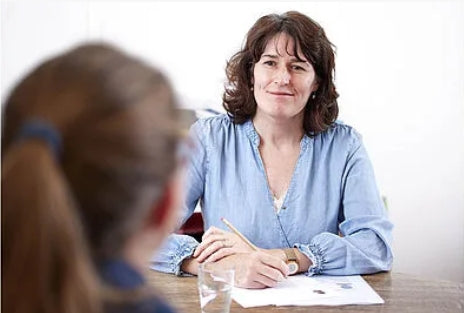 Eine Frau in einer hellblauen Bluse sitzt an einem Tisch und hält einen Stift in der Hand. Vor ihr liegen Papiere sowie ein Glas Wasser. Im Vordergrund ist der Rücken einer anderen Person zu sehen, die ihr gegenüber sitzt."