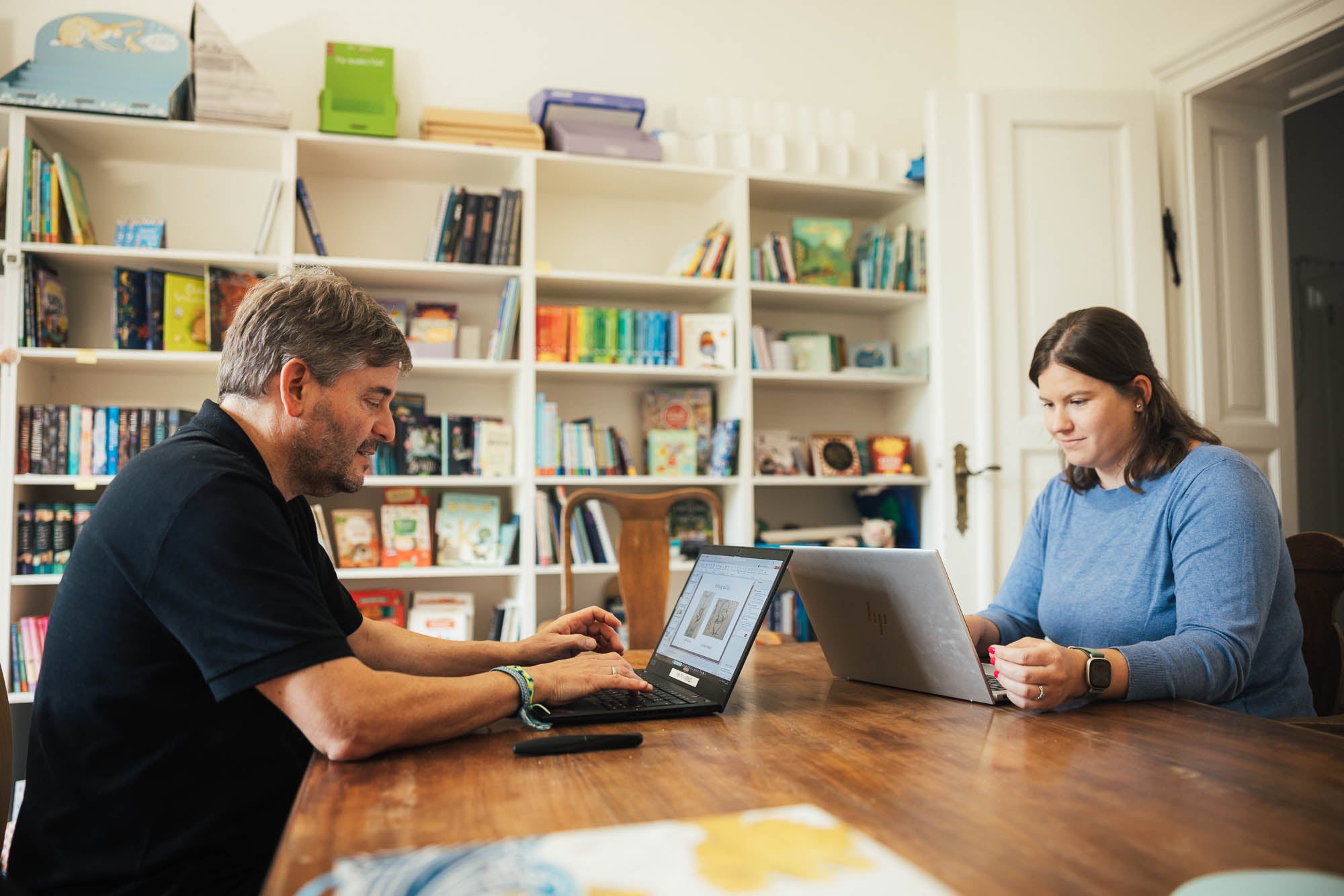 Zwei Personen sitzen an einem Holztisch in einem Raum mit Bücherregalen, die farbenfrohe Buchrücken und Deko-Elemente enthalten. Beide arbeiten an Laptops und scheinen in einem geschäftlichen Gespräch oder einer Zusammenarbeit vertieft zu sein. Die Atmosphäre ist kreativ und professionell mit einem Fokus auf Büchermaterialien.