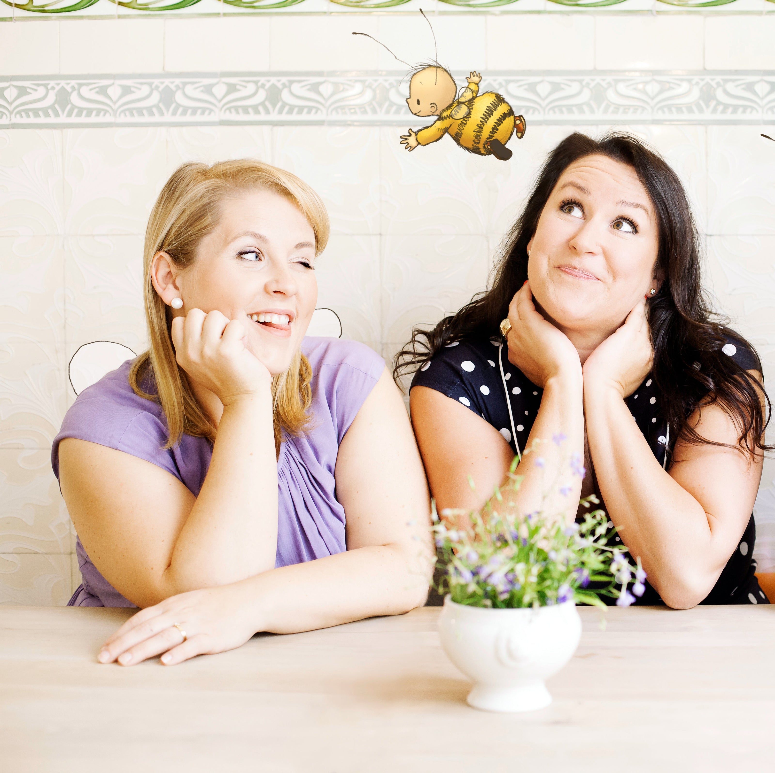 Zwei Frauen, Maite Kelly und Britta Sabbag, sitzen an einem hellen Holztisch. Vor ihnen steht eine kleine weiße Vase mit Lavendel. Im Hintergrund ist eine dekorative Fliesenwand zu sehen. Oberhalb der Szene schwebt die Zeichnung der Figur "Die kleine Hummel Bommel", die aus ihrem gemeinsamen Buch stammt.