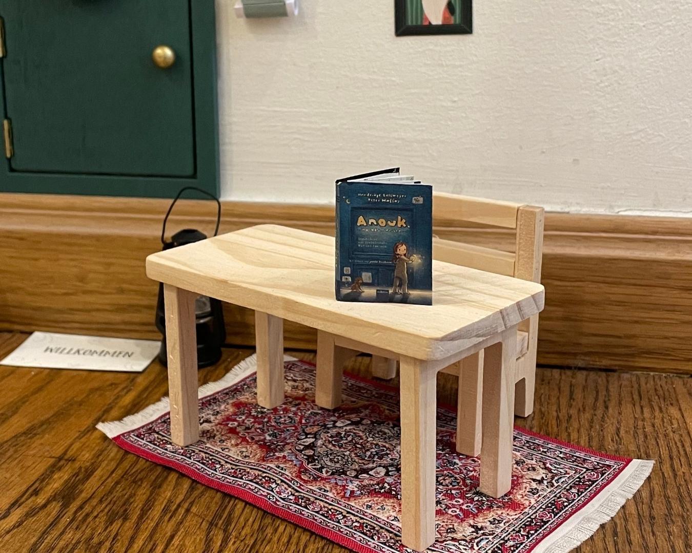 Miniaturszene mit einem kleinen Holztisch und einer passenden Holzbank, die auf einem detailreichen, orientalischen Teppich steht. Auf dem Tisch liegt ein aufgeschlagenes Buch mit dem Titel 'Anouk'. Im Hintergrund sind eine grüne Tür mit goldfarbenem Griff, ein Bilderrahmen und ein schwarzes Objekt zu sehen. Die Szene ist liebevoll gestaltet und wirkt wie eine winzige Wohnumgebung."