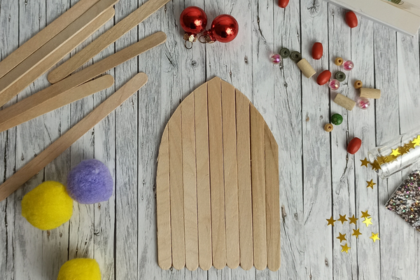 Bastelszene mit einer Miniatur-Wichteltür aus Holzstäbchen, arrangiert auf einer hellen Holzoberfläche. Um die Tür herum liegen bunte Filzkugeln, rote Weihnachtskugeln, Holzperlen, Glitzersterne und weitere Bastelmaterialien. Die Szene vermittelt eine kreative und festliche Atmosphäre.