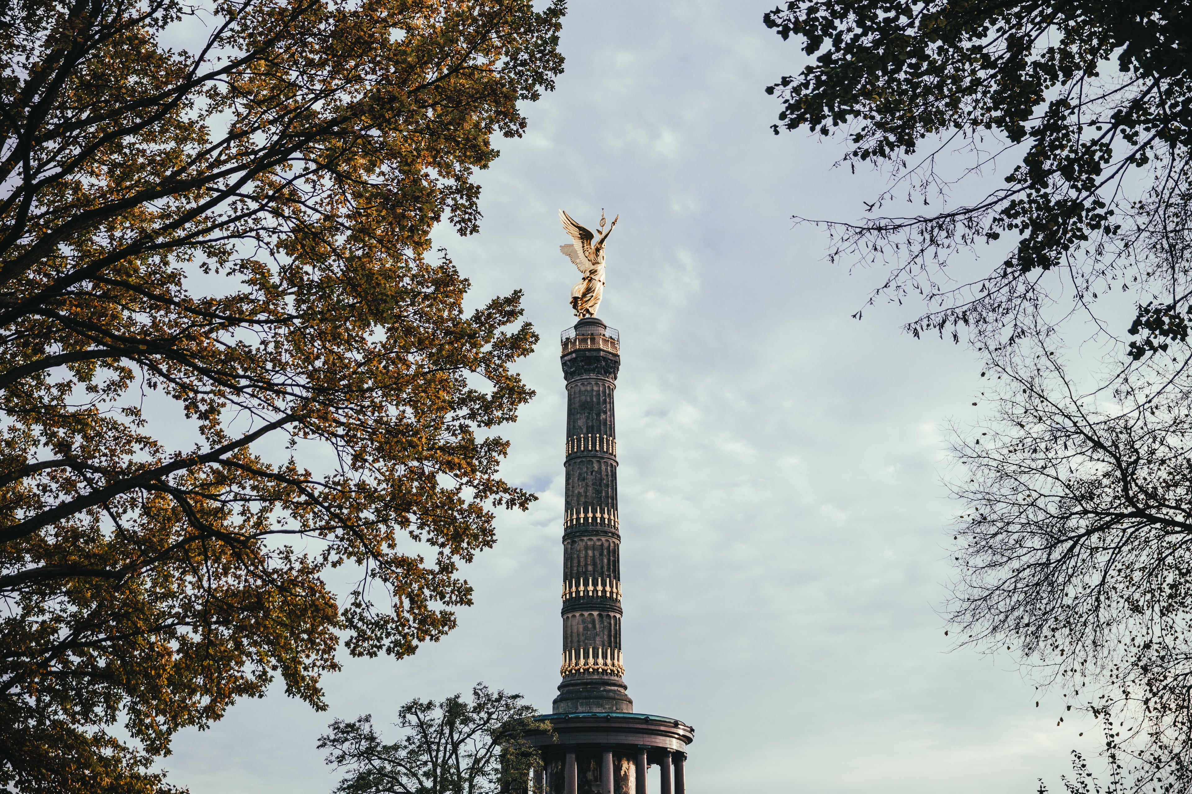 Die Berliner Siegessäule mit der vergoldeten Viktoria-Statue an der Spitze, umgeben von herbstlichen Bäumen. Der Himmel ist leicht bewölkt und verleiht der Szene eine ruhige Atmosphäre.