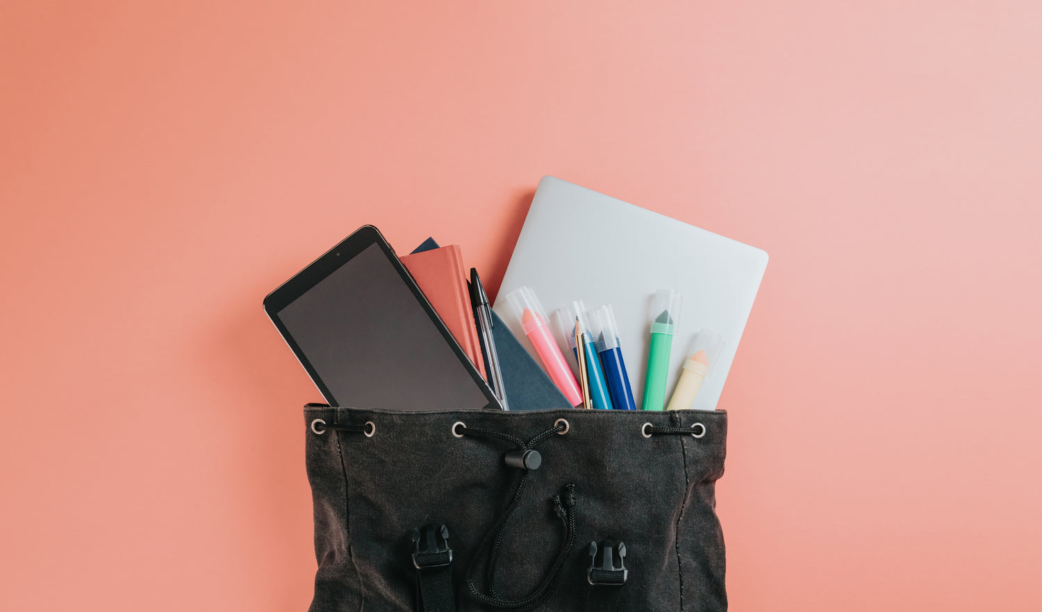 Offener schwarzer Rucksack vor einem rosa Hintergrund. Herausstehend ein Tablet, Bücher, ein Laptop, Stifte und Textmarker in verschiedenen Farben.