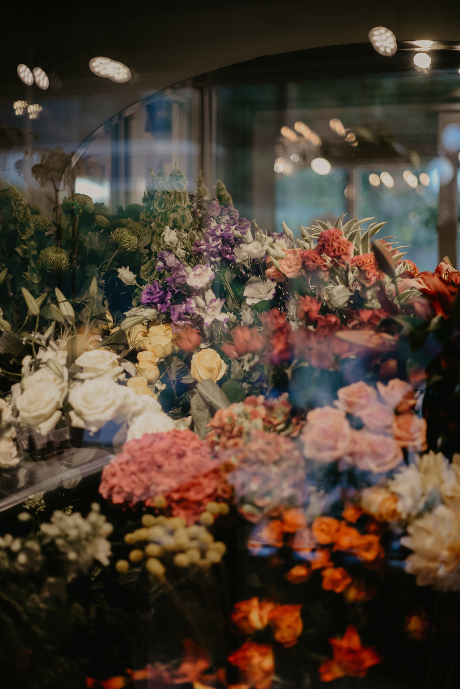 Eine bunte Auswahl an Blumen in einem Schaufenster, darunter Rosen, Hortensien, Lilien und andere Blüten in verschiedenen Farben wie Rot, Weiß, Rosa, Lila und Orange. Die Reflexionen im Glas zeigen Lichter und einen verschwommenen Hintergrund.