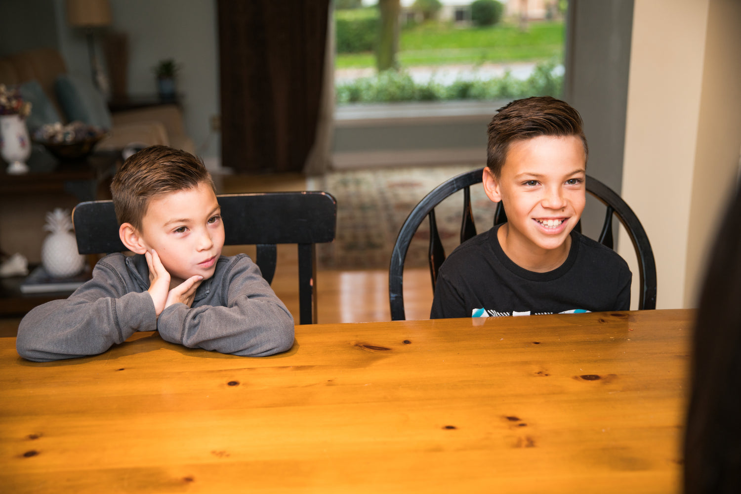 Zwei Kinder sitzen an einem Holztisch in einem gemütlichen Wohnraum. Im Hintergrund sind ein Sofa mit Kissen, Dekorationen und ein großes Fenster mit Blick auf eine grüne Außenlandschaft zu sehen.