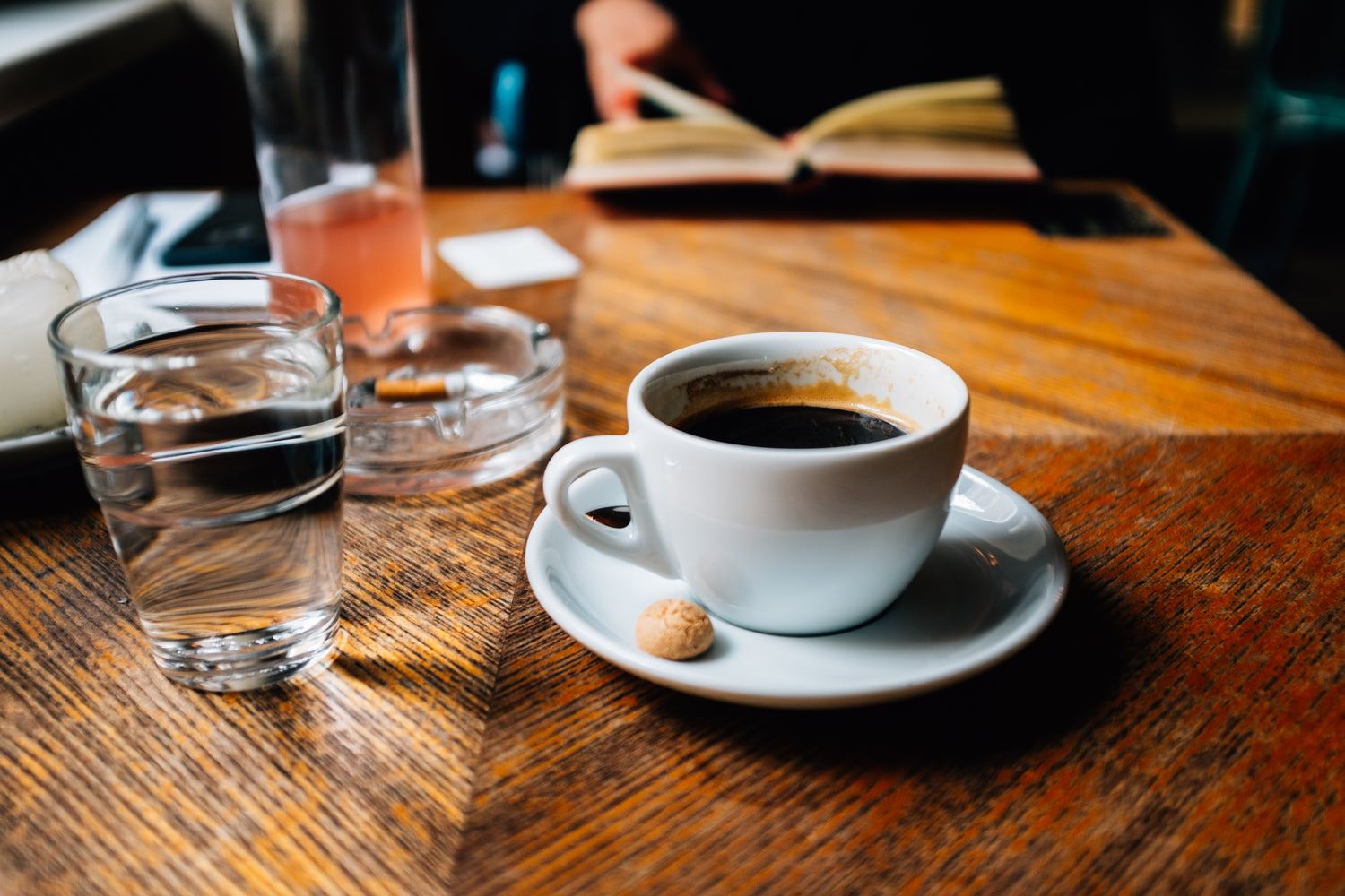 Eine weiße Tasse mit schwarzem Kaffee auf einer Untertasse, daneben ein kleines Gebäck, steht auf einem Holztisch. Im Hintergrund ein Glas Wasser, ein Aschenbecher und eine Person, die ein Buch liest.