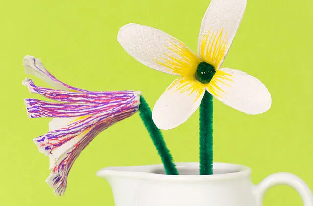 Zwei handgefertigte Blumen in einer kleinen weißen Vase auf einem hellgrünen Hintergrund. Eine Blume mit weißen Blütenblättern und gelbem Zentrum, die andere in Lila-Tönen.