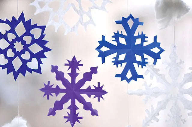Bunte Papier-Schneeflocken in Blau und Lila, aufgehängt vor einem hellen, unscharfen Hintergrund.